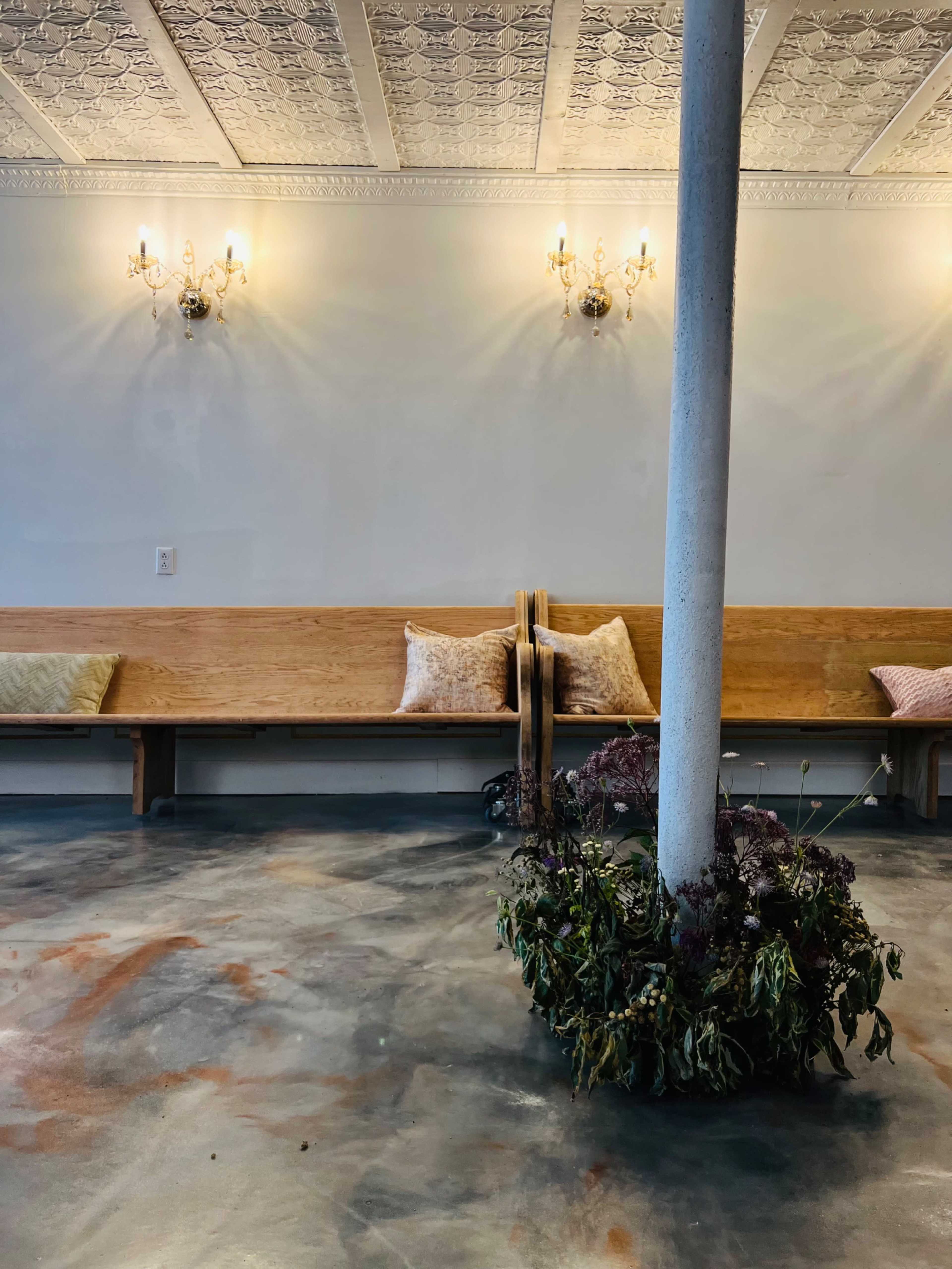 A minimalistic interior features wooden benches with cushions, a central column, and a planter with dried foliage on a polished concrete floor.