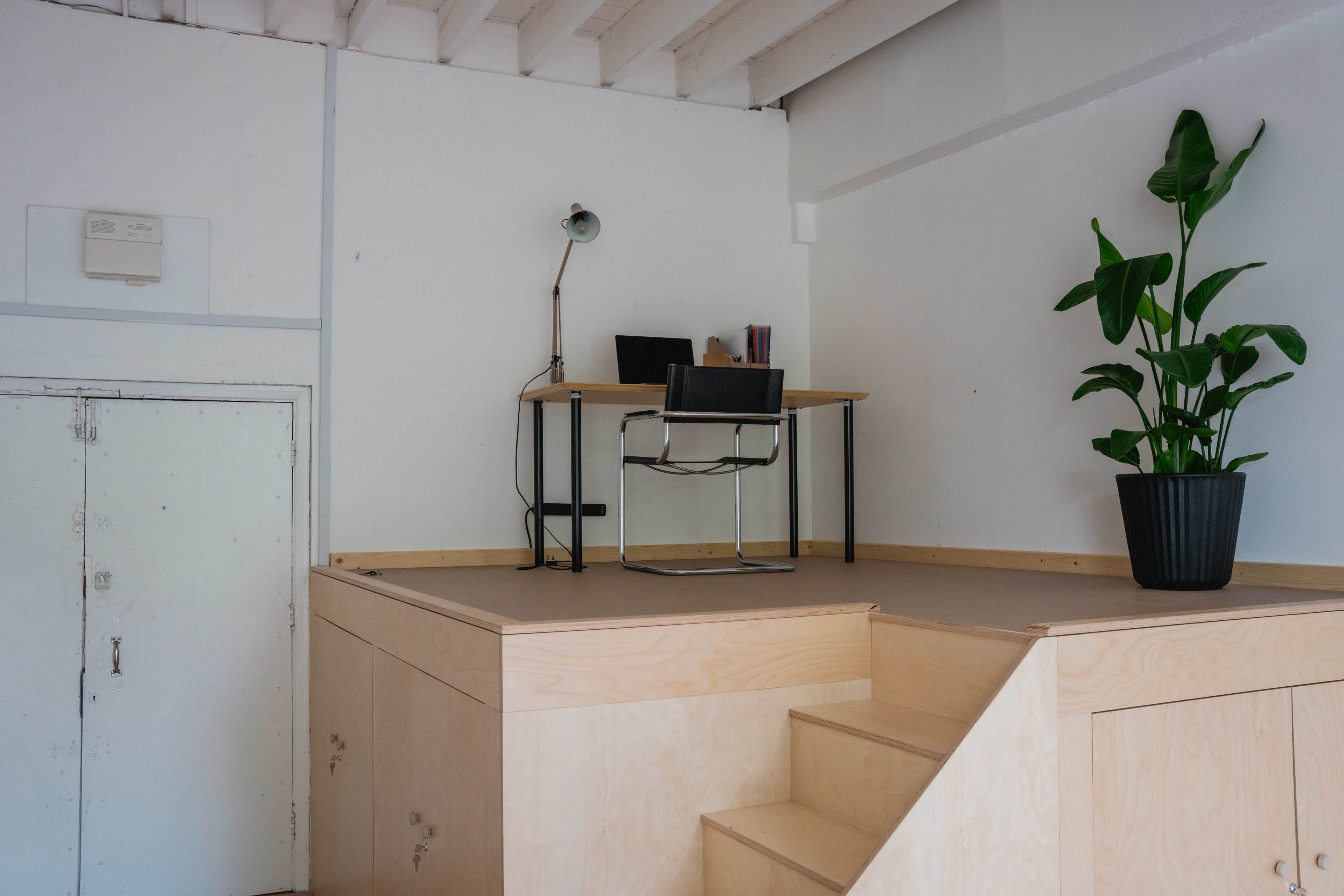 A minimalist workspace features a desk and a chair positioned on a raised platform, with a potted plant nearby and stairs leading up to it.