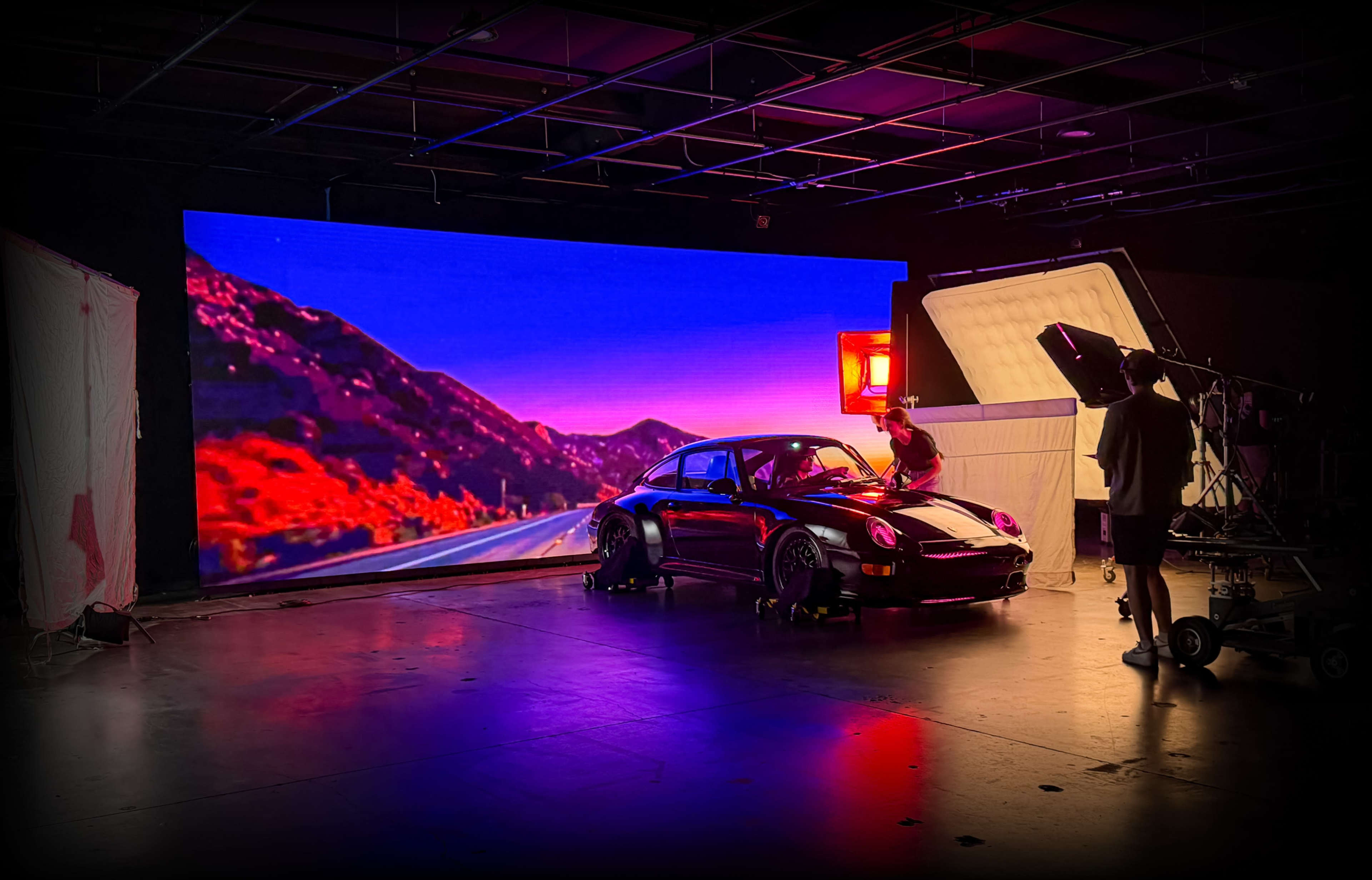 A black car is positioned in a studio setting with a large screen displaying a colorful mountain landscape in the background.