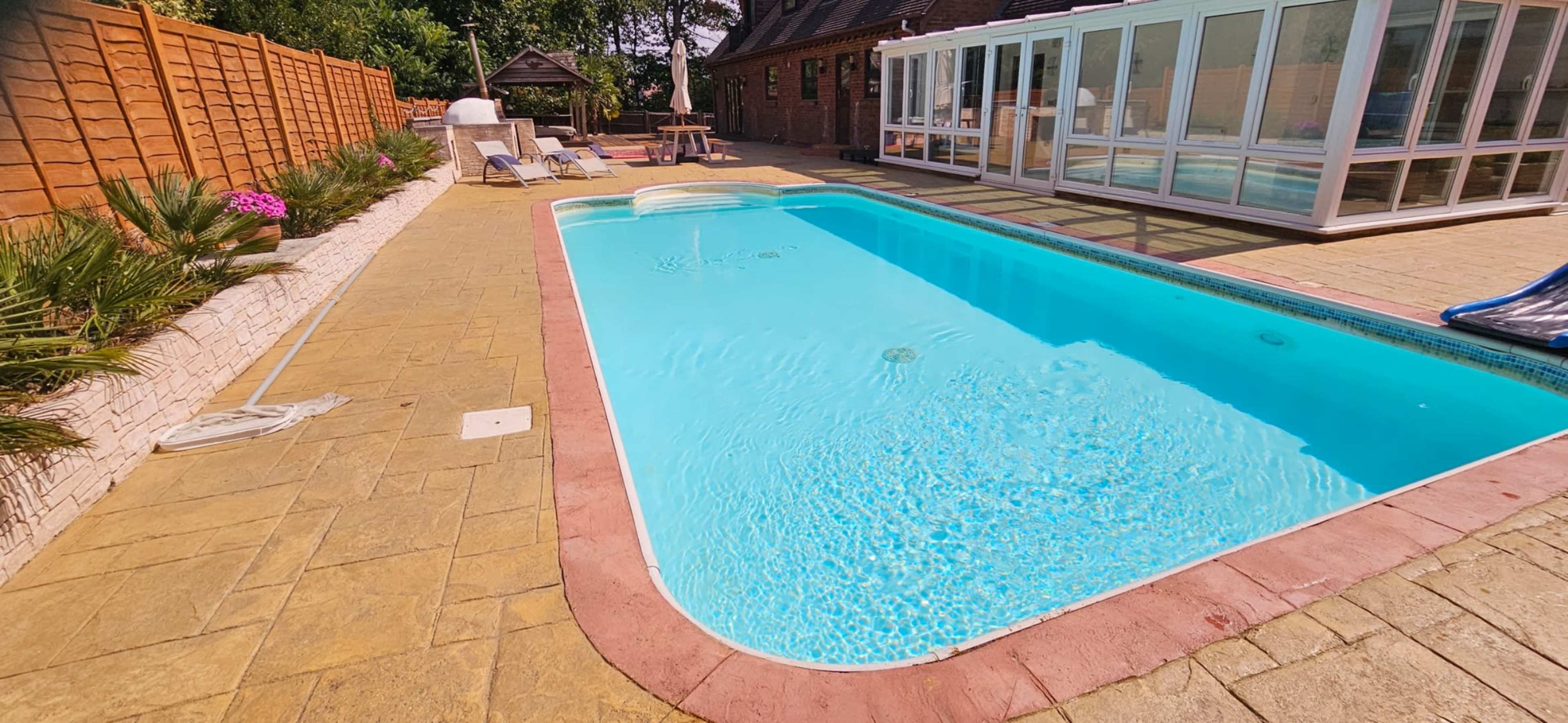 The image shows a rectangular swimming pool surrounded by a stone patio, chaise lounges, and a glass-enclosed sunroom in a residential backyard.