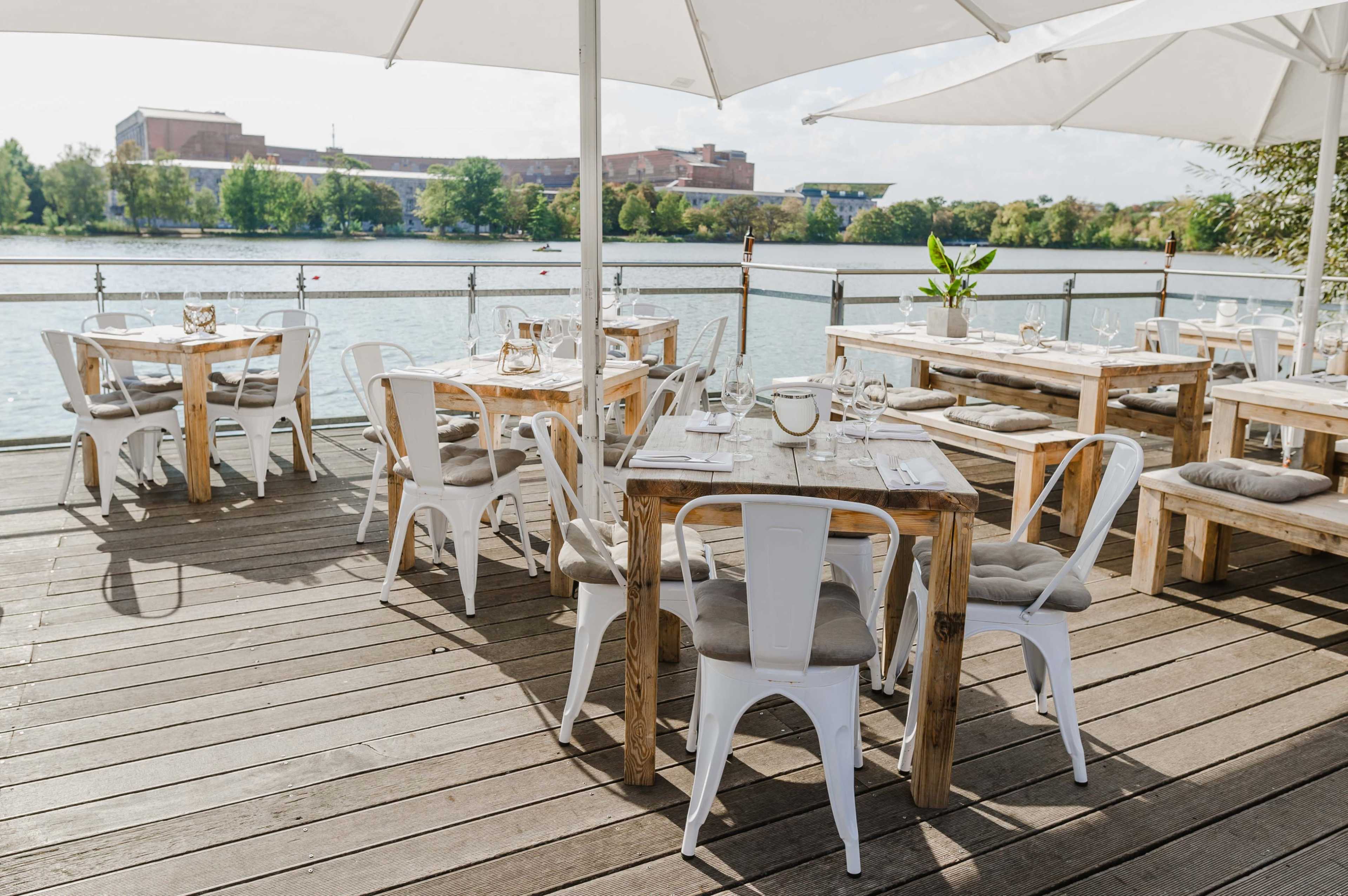 The image shows an outdoor dining area with wooden tables and white chairs set up along a waterfront.
