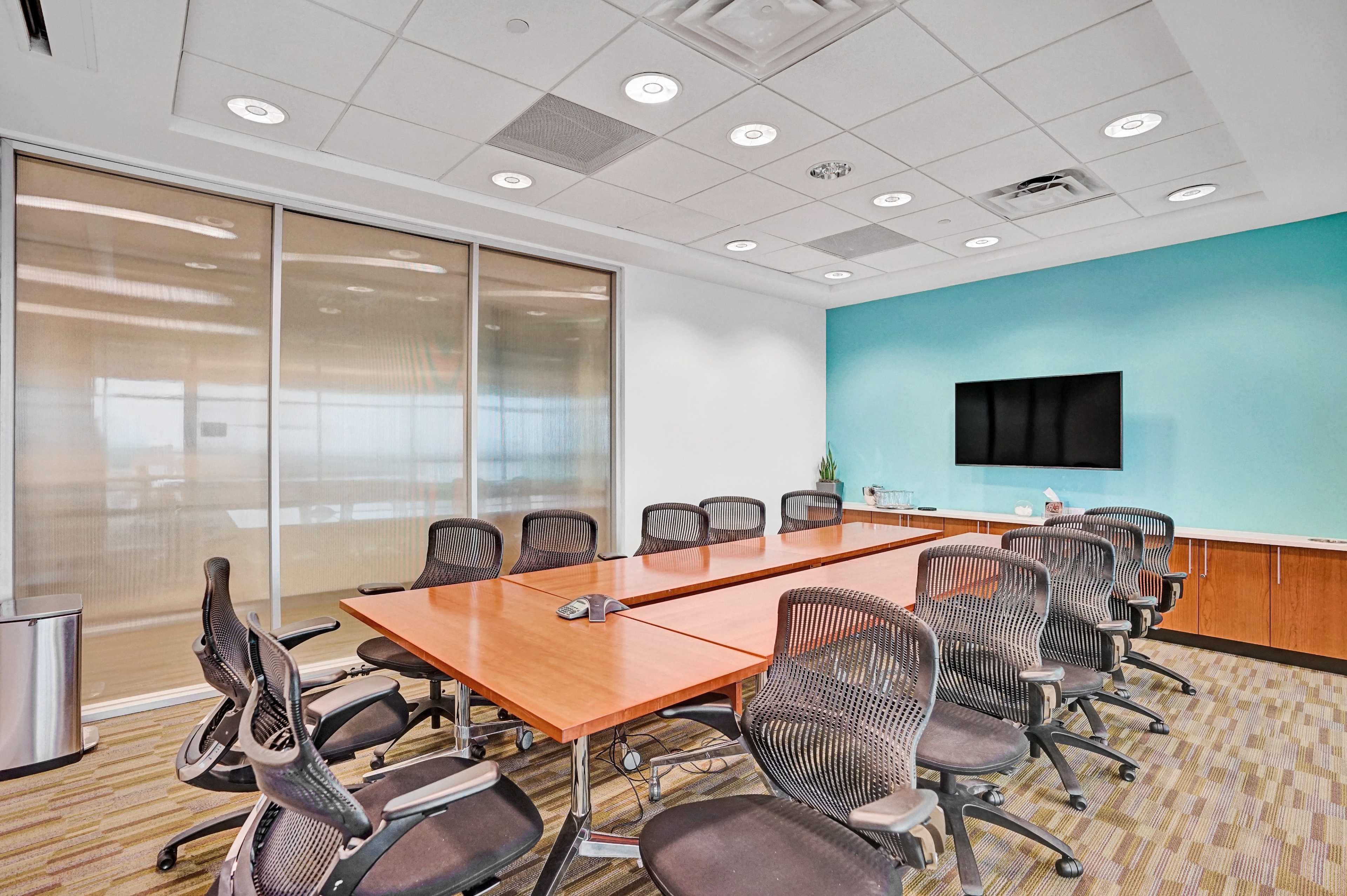 A modern conference room features a long wooden table surrounded by ergonomic chairs, with a large screen mounted on a turquoise wall and glass partitions allowing natural light.