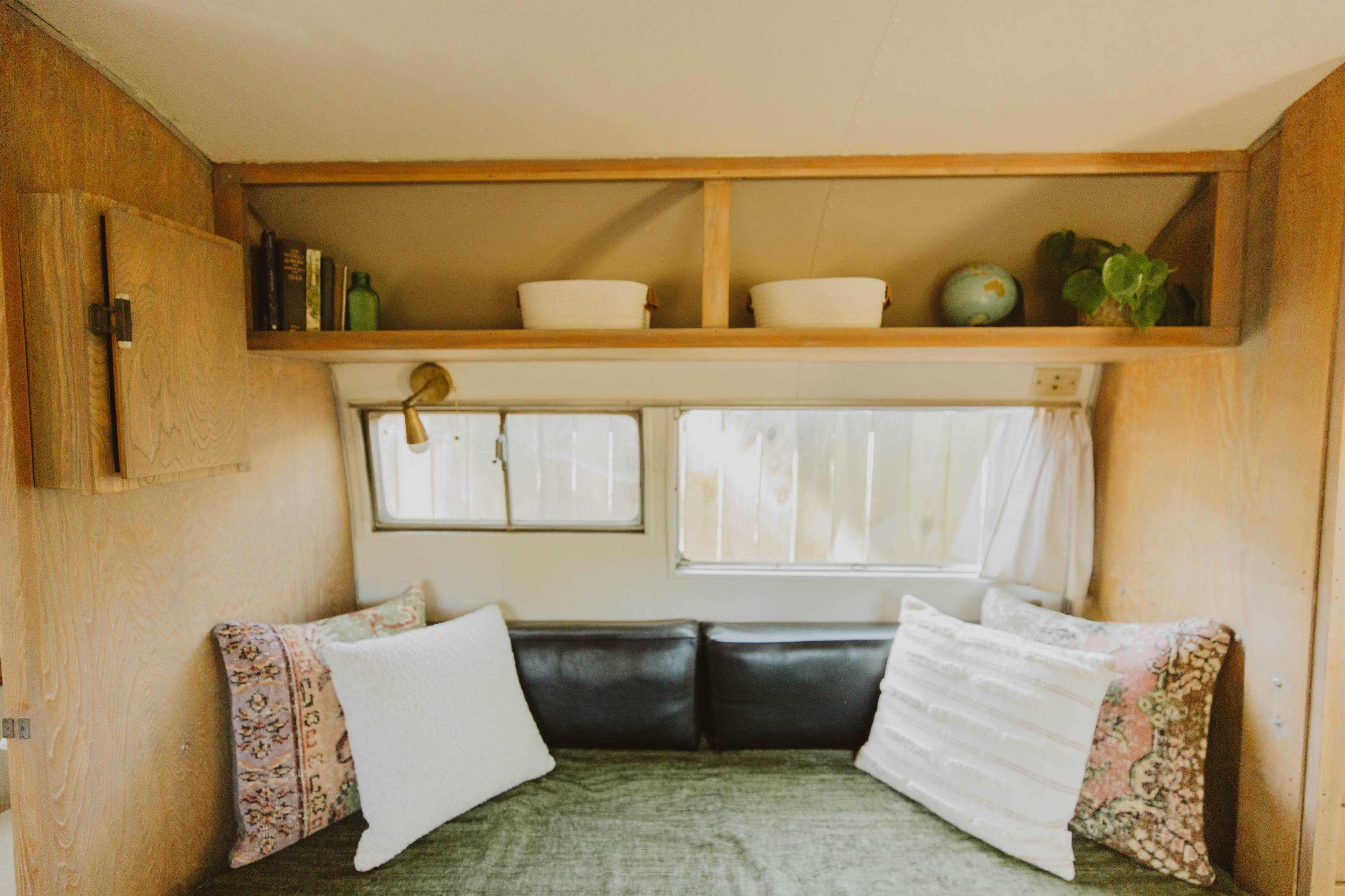 A cozy interior of a camper with a black sofa, decorative pillows, and shelves above featuring books and plants.