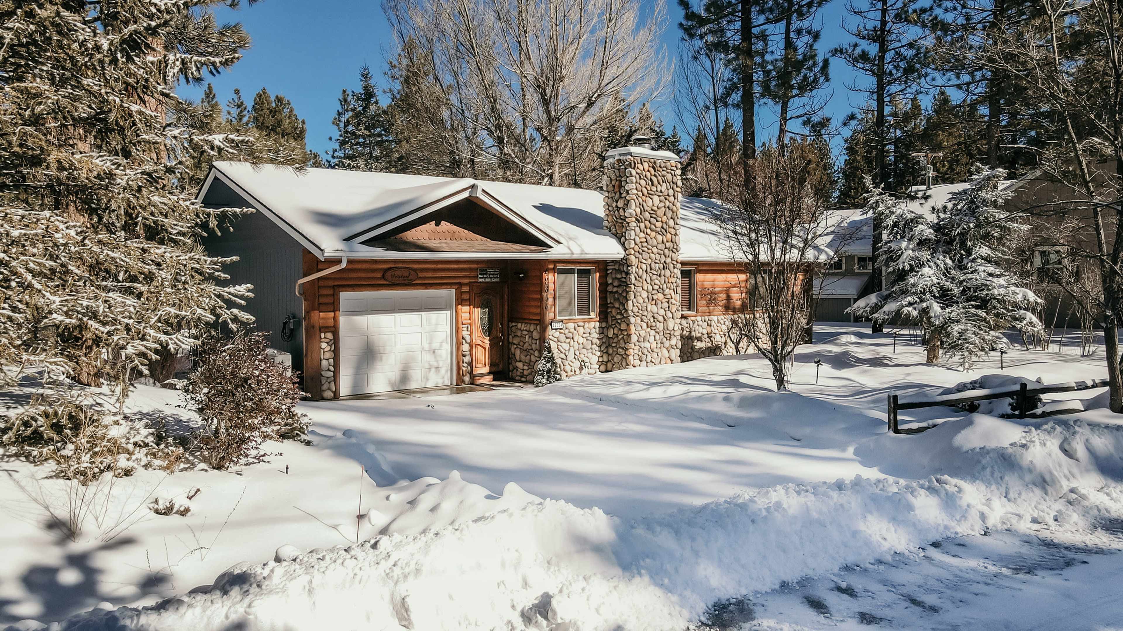 A single-story house with a stone chimney is surrounded by snow-covered trees and a white landscape.