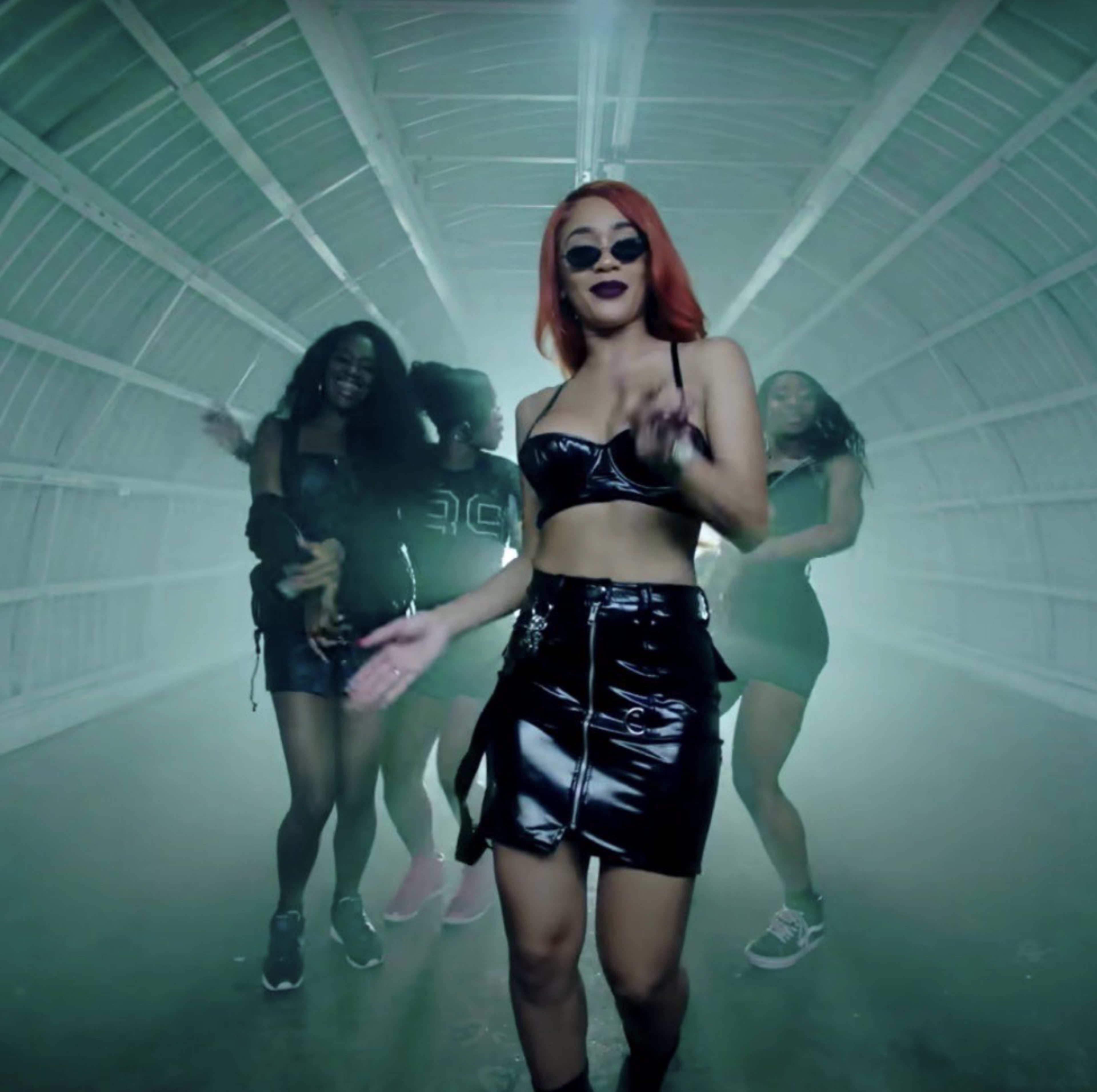 A group of four women, dressed in stylish outfits, dances in a dimly lit, industrial space with a foggy atmosphere.
