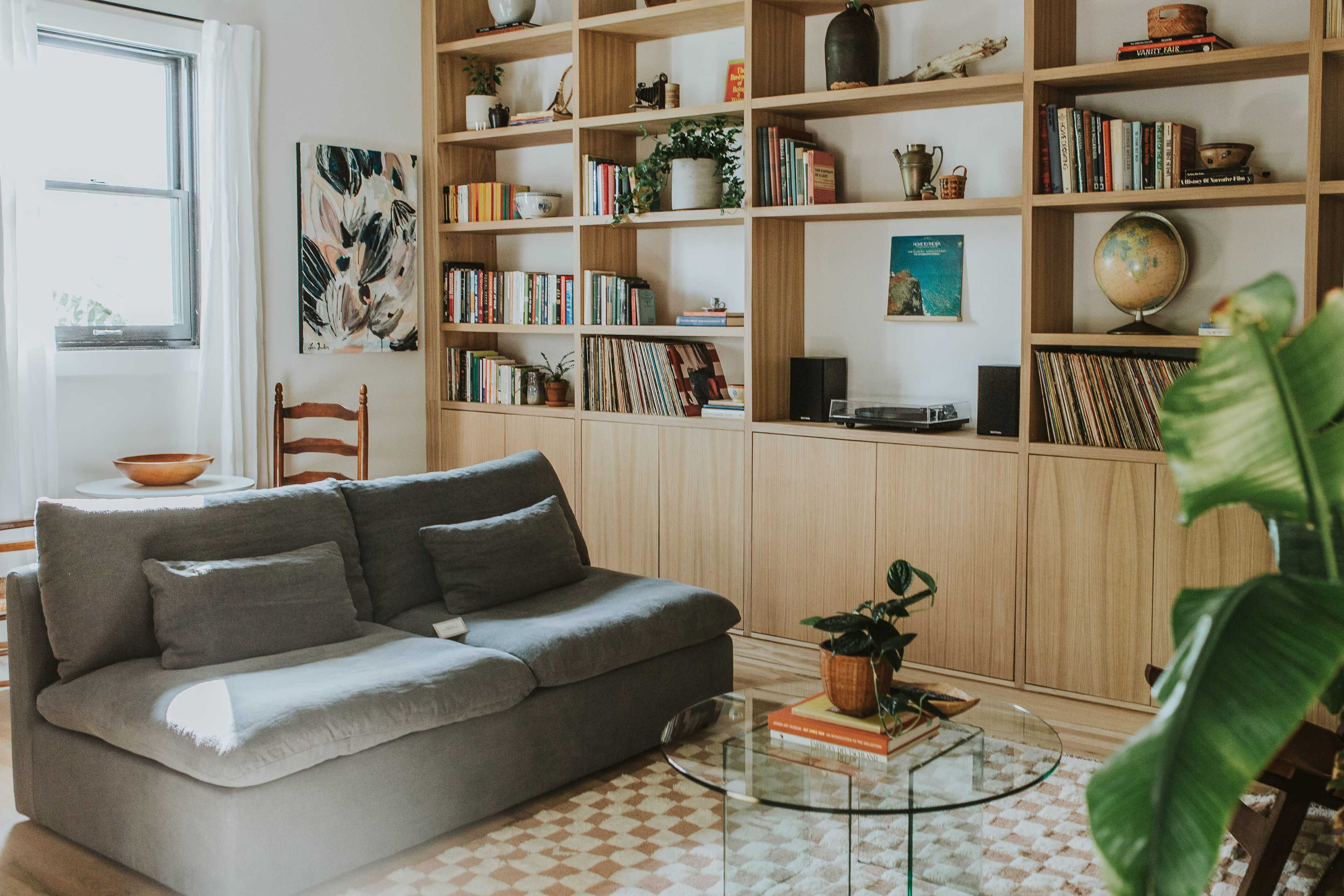 A cozy living room features a gray sofa, a glass coffee table, and wooden shelves filled with books and decorative items.