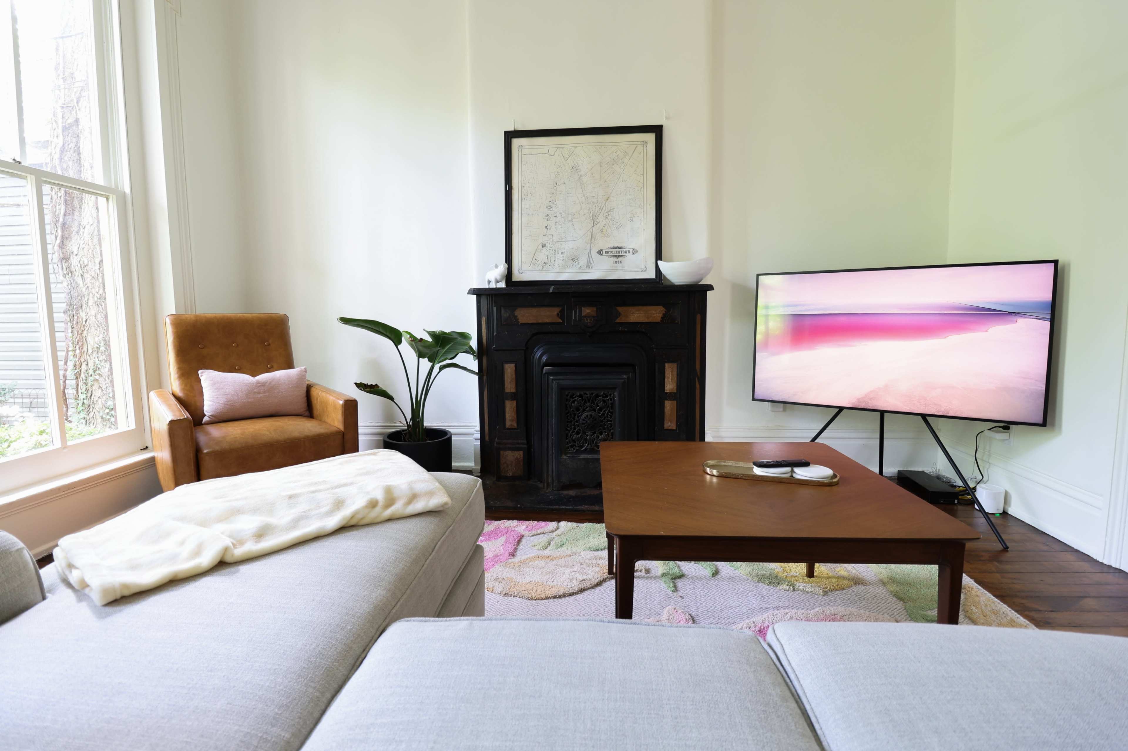 A living room features a brown armchair, a modern television on a stand, a wooden coffee table, a plant, and a decorative fireplace.