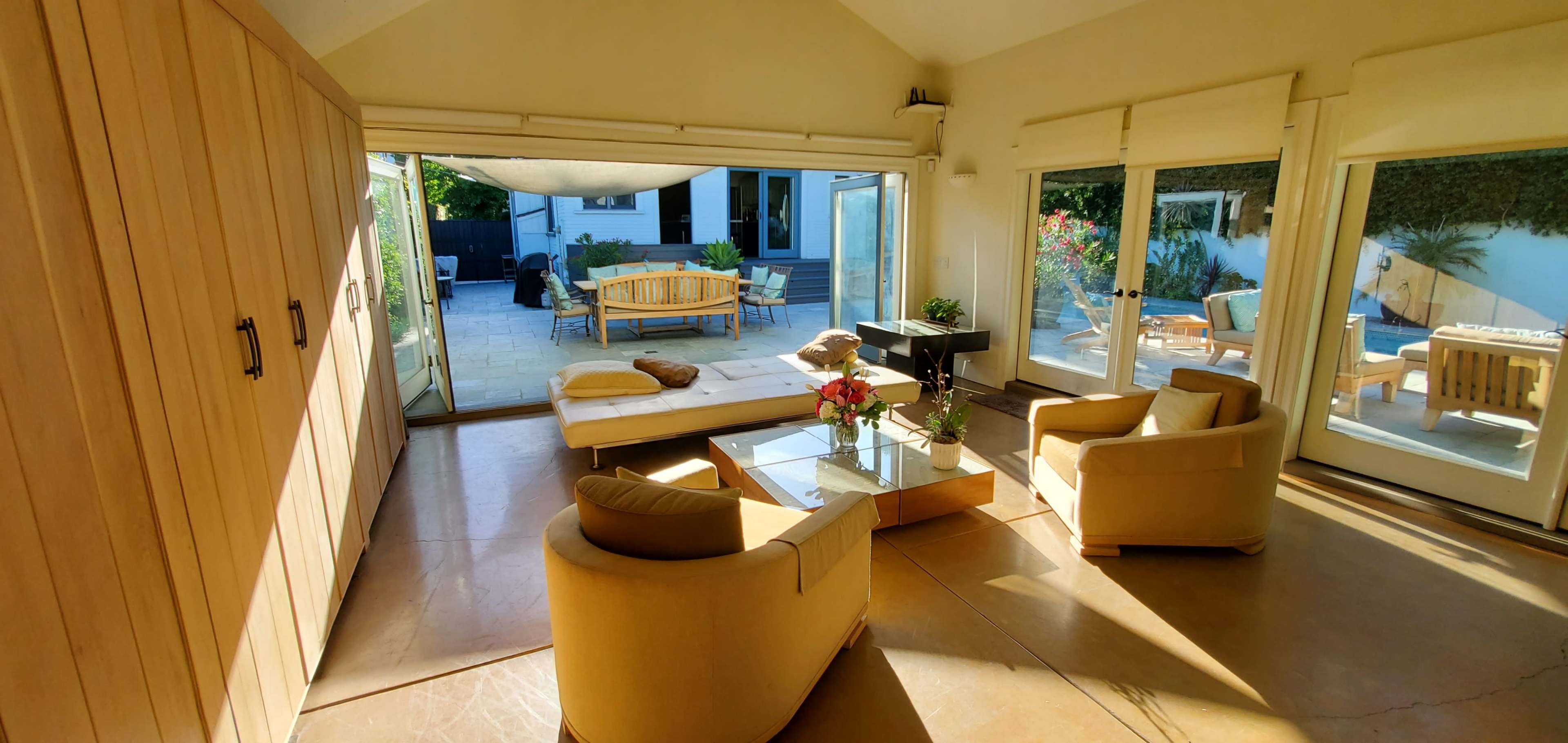 The image shows a sunlit room with a modern seating area, large windows opening to an outdoor patio with additional seating.