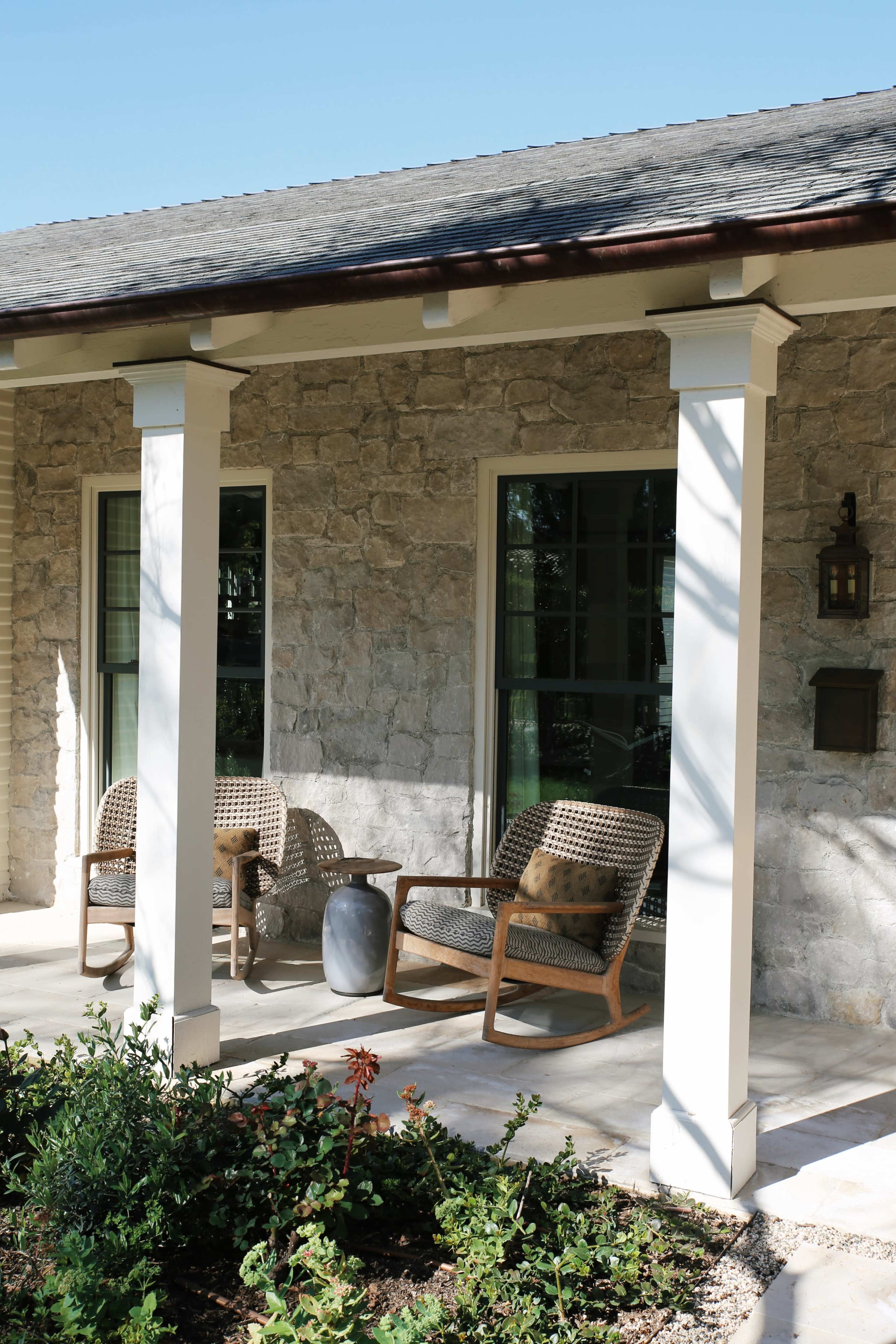 A stone cottage porch with two woven chairs, a small side table, and a lantern mounted on the wall.