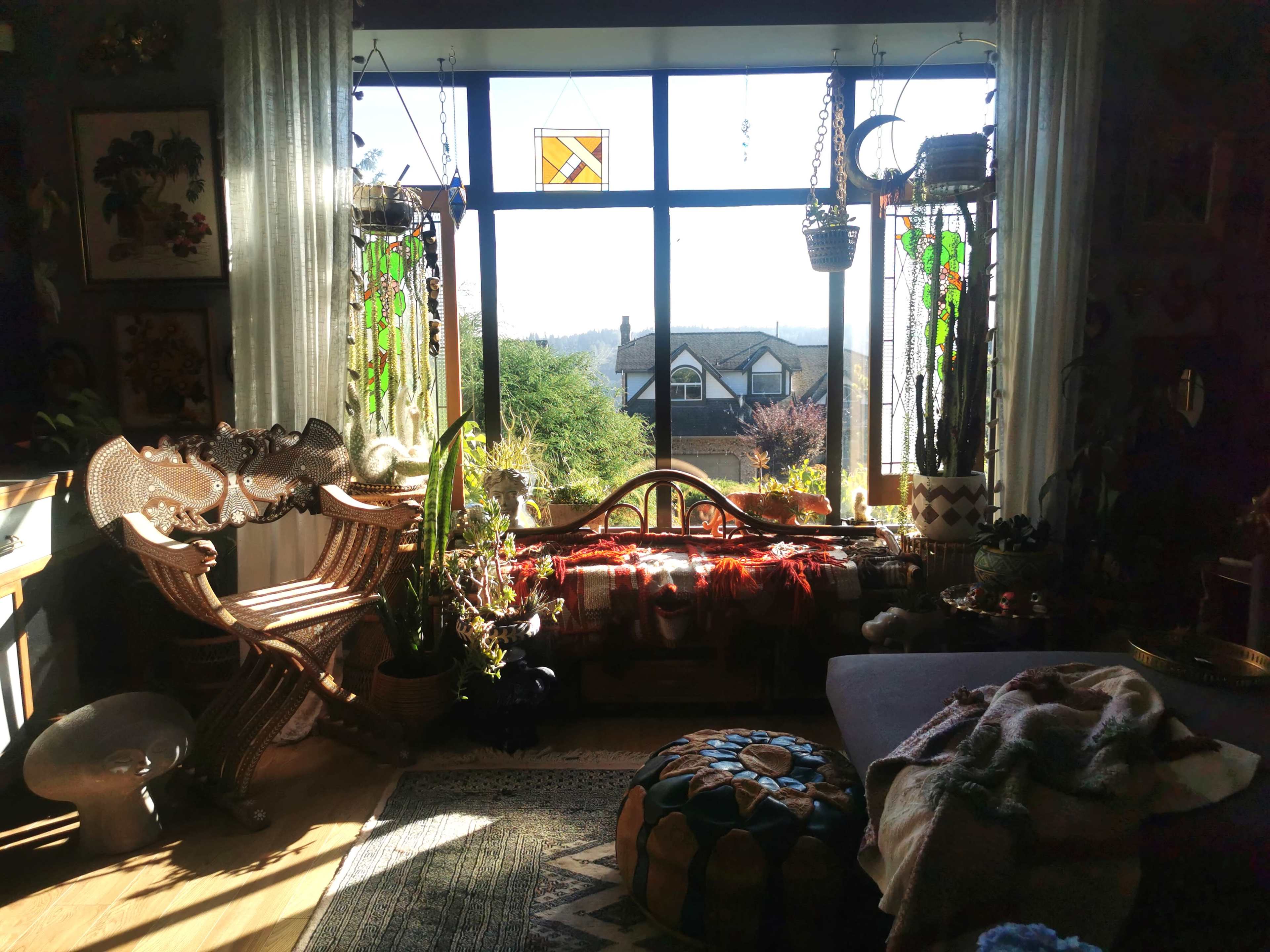 The room features a large window adorned with stained glass, offering a view of greenery and a house in the distance, while plants and decorative items are arranged around a wooden bench and a cozy seating area.