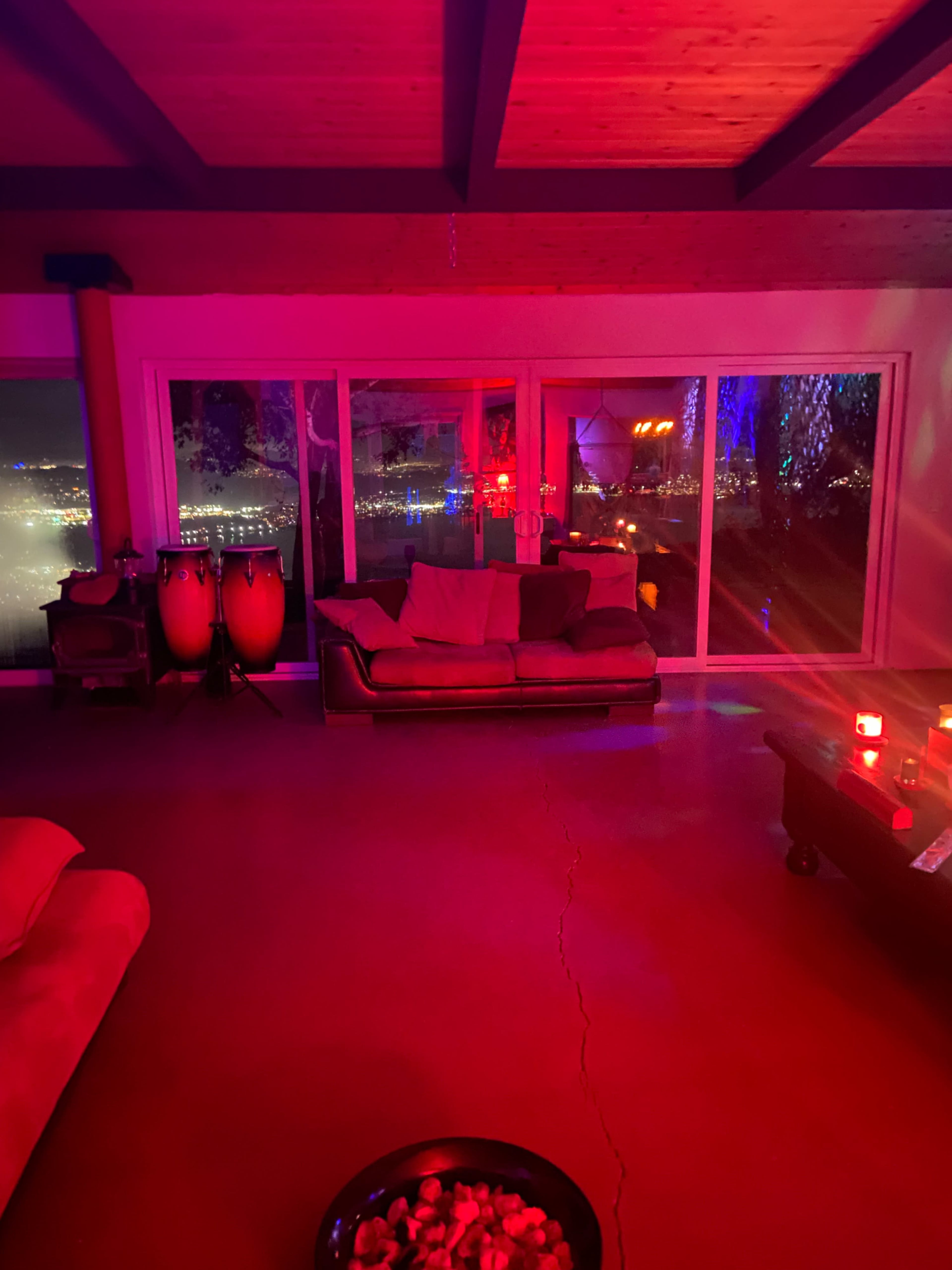 The image shows a dimly lit living room with a large couch, a coffee table, and colorful lighting, overlooking a city skyline through large glass windows.