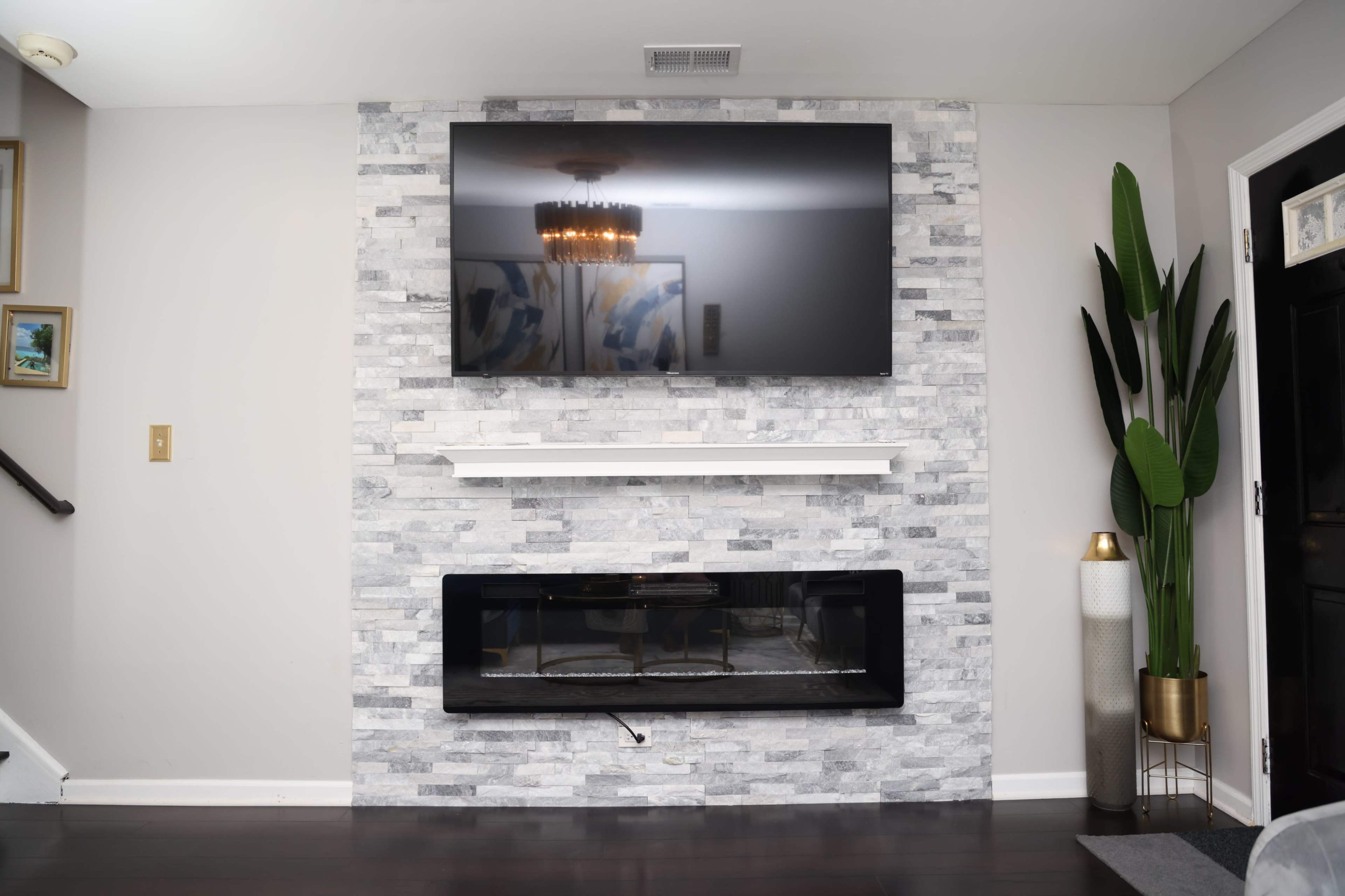 A modern living room with a mounted flat-screen television above a stone fireplace, flanked by a tall plant in a decorative pot.