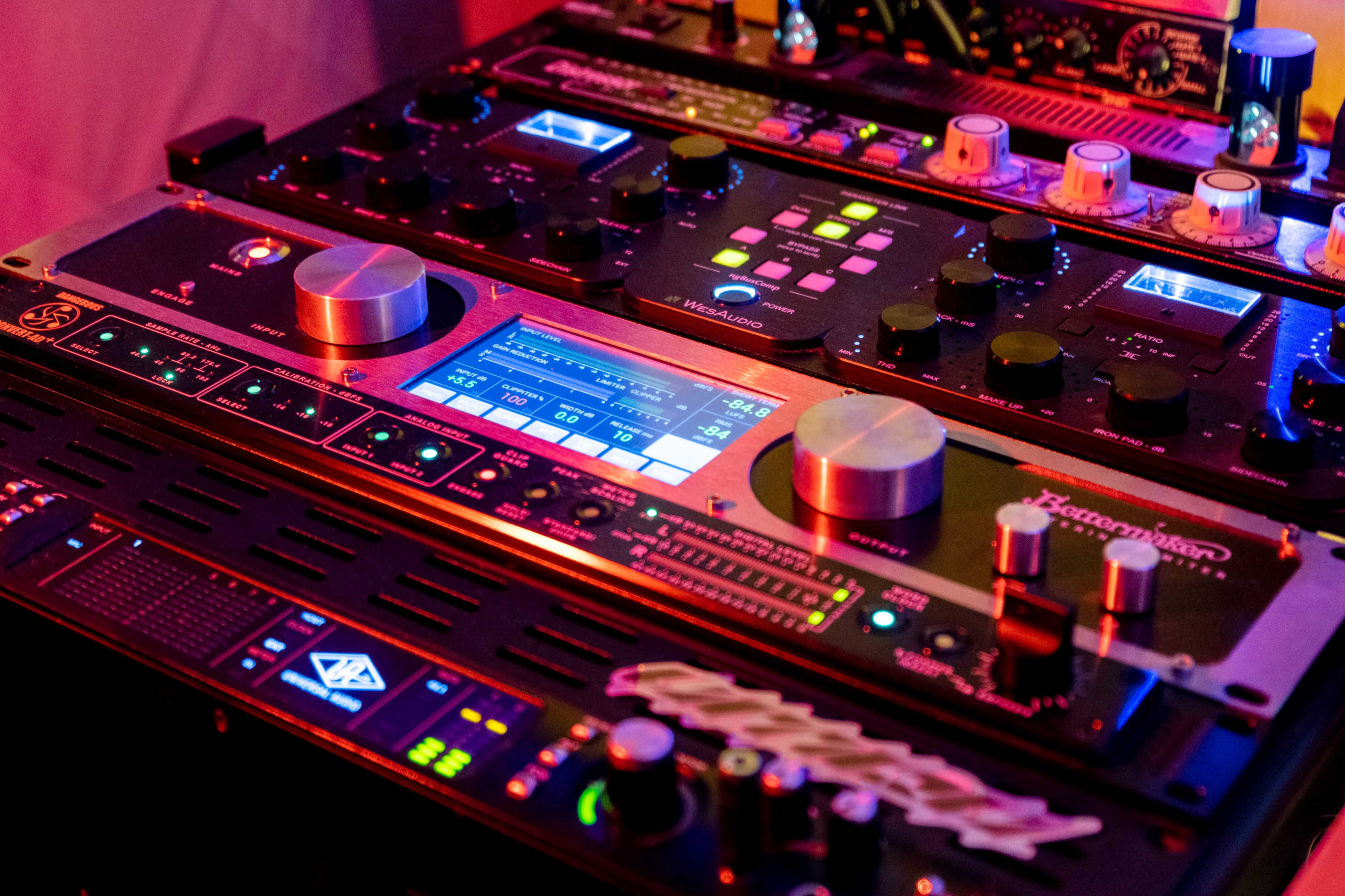 The image shows a close-up of a complex array of audio mixing equipment featuring various knobs, sliders, and digital displays illuminated by colorful lights.