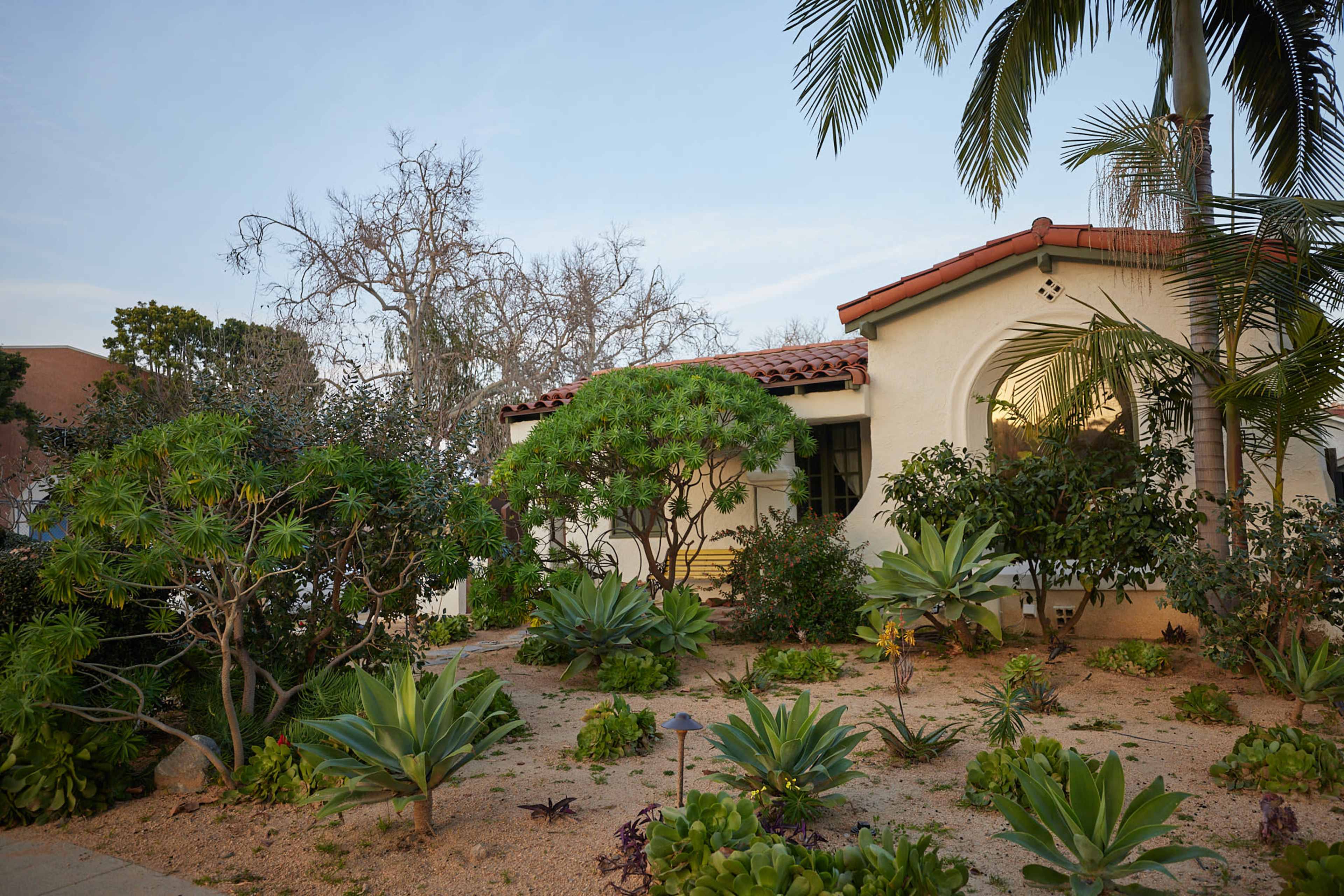 A white stucco house with a red-tiled roof is surrounded by a garden featuring various plants and shrubs.