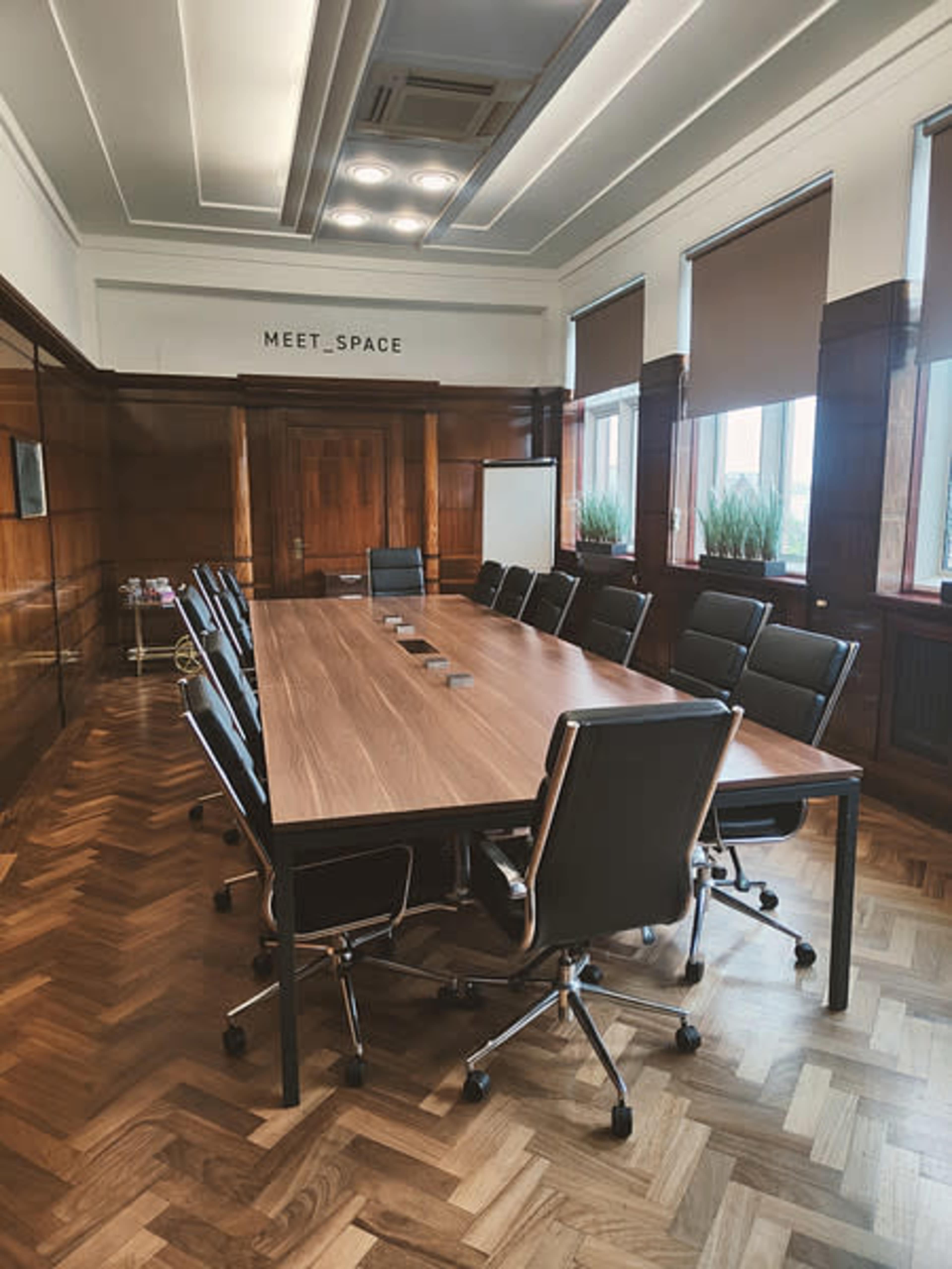 A long wooden conference table with black swivel chairs is arranged in a well-lit meeting room featuring wooden paneling and large windows.
