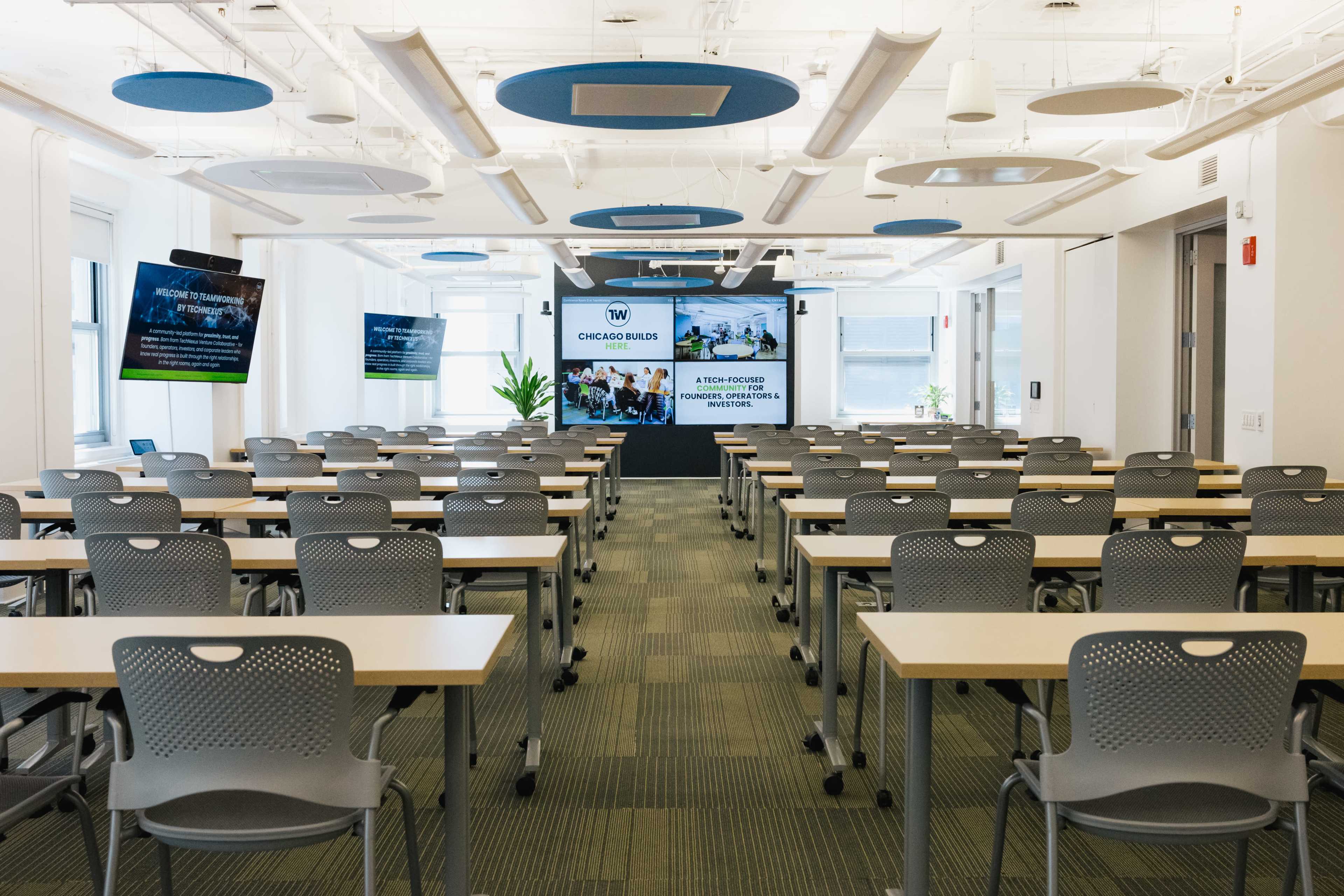 The image shows a spacious conference room set up with rows of tables and chairs, equipped with a screen and presentation area at the front.