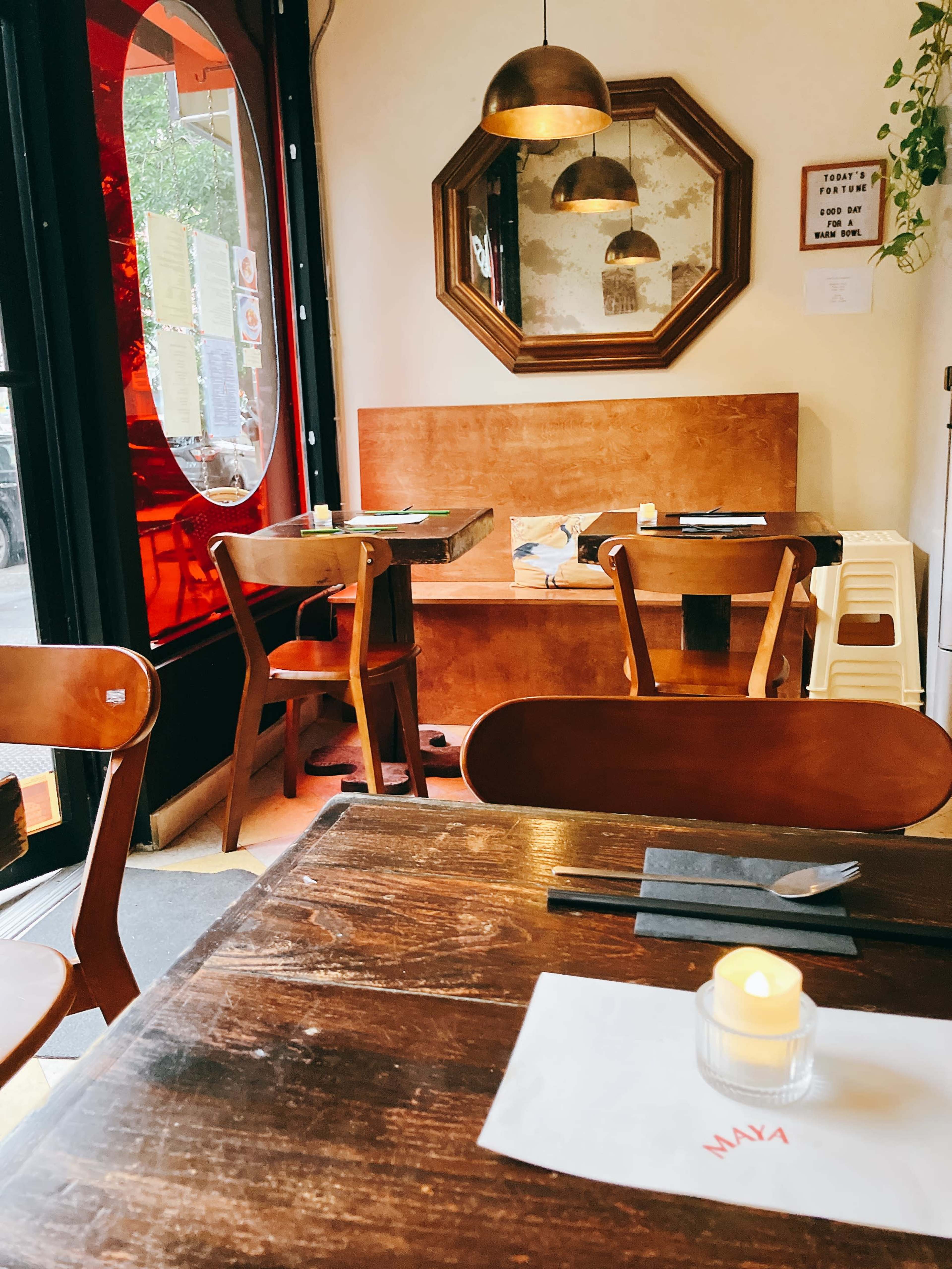 The image shows a cozy restaurant interior with wooden tables, chairs, and a large mirror reflecting the space, while a candle sits on a table in the foreground.