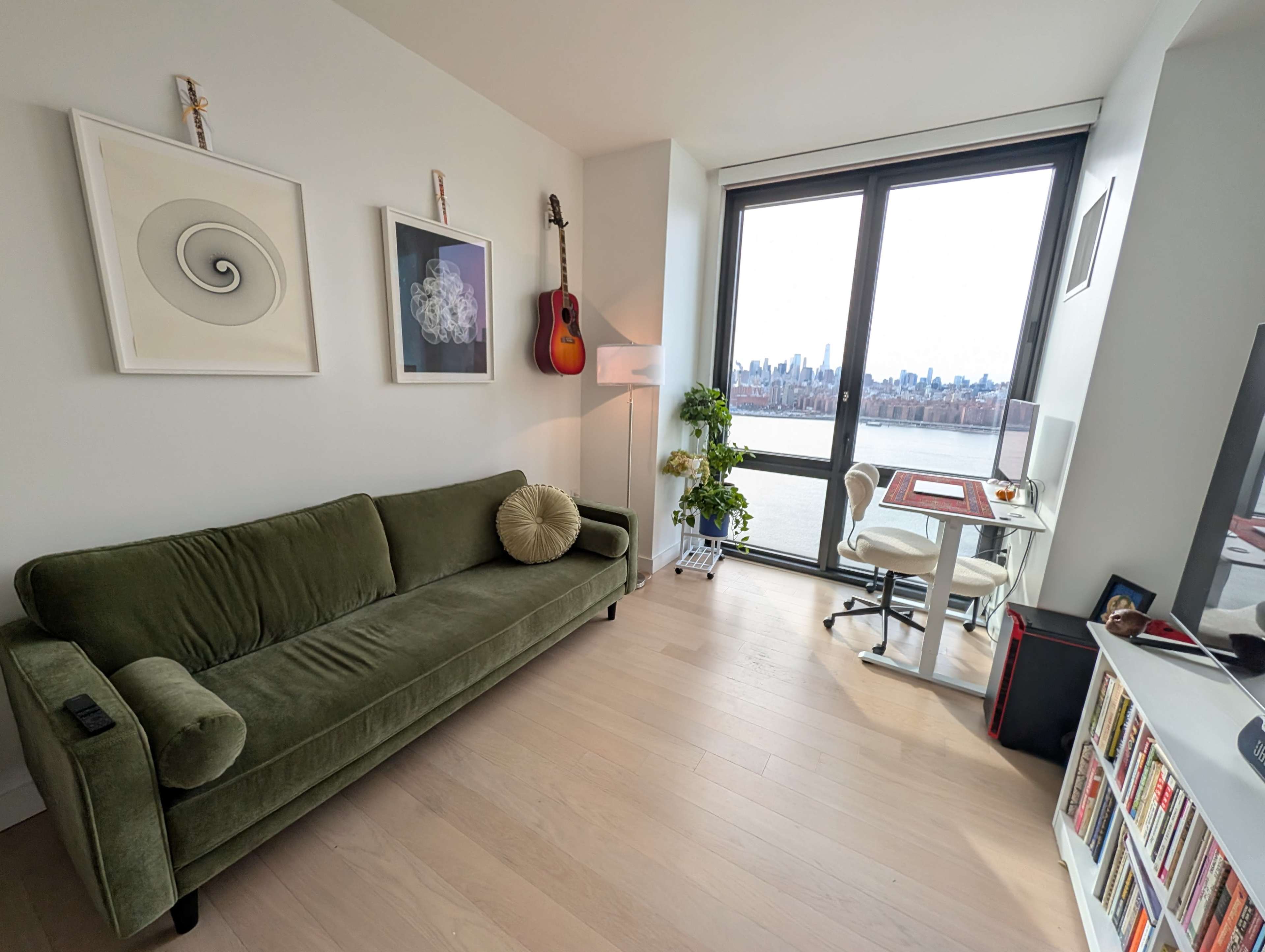 The image shows a modern living space featuring a green sofa, a guitar on the wall, a desk with a chair by a large window overlooking a city skyline.