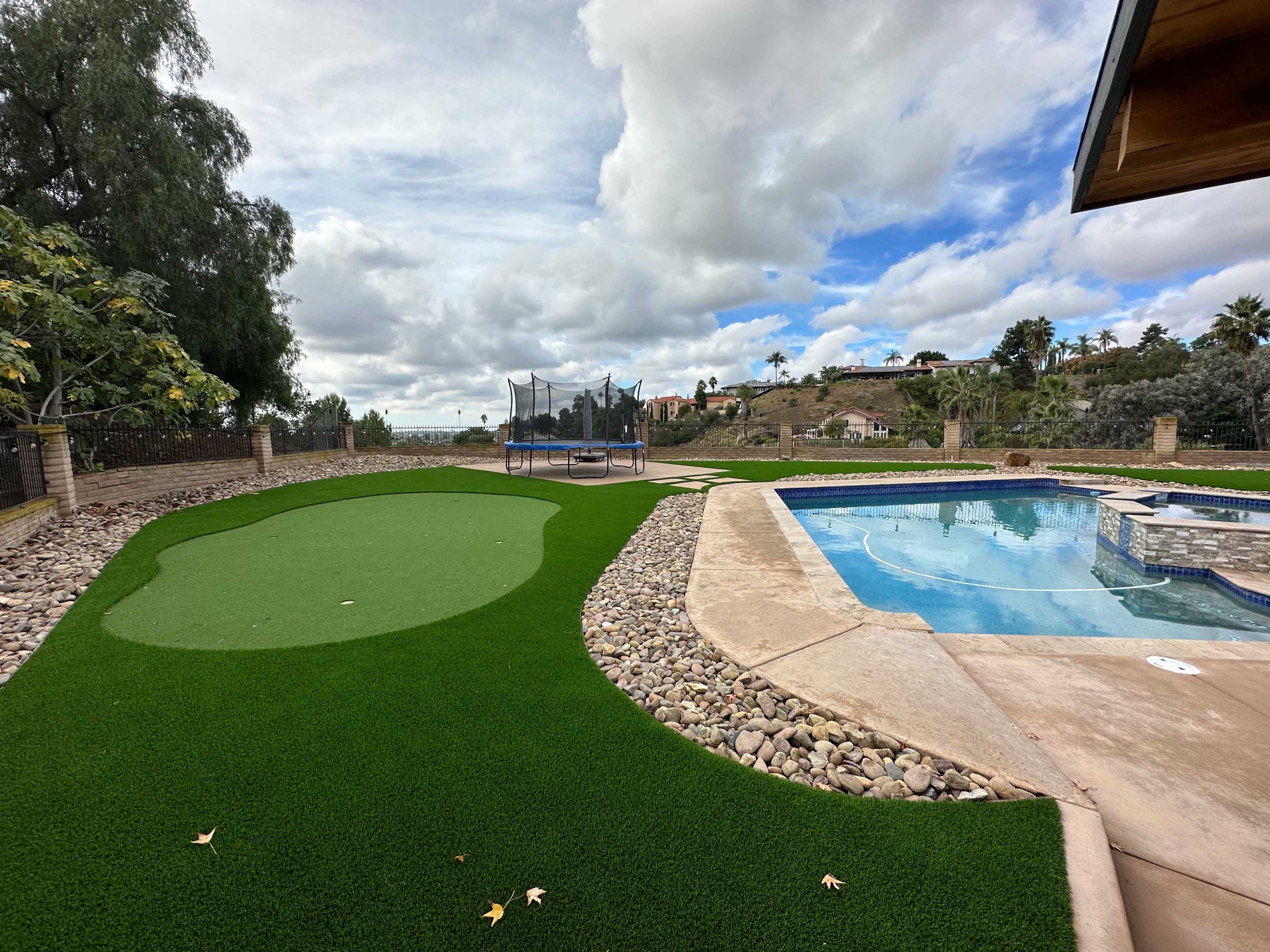 A backyard features a putting green alongside a pool and a trampoline in the distance under a cloudy sky.