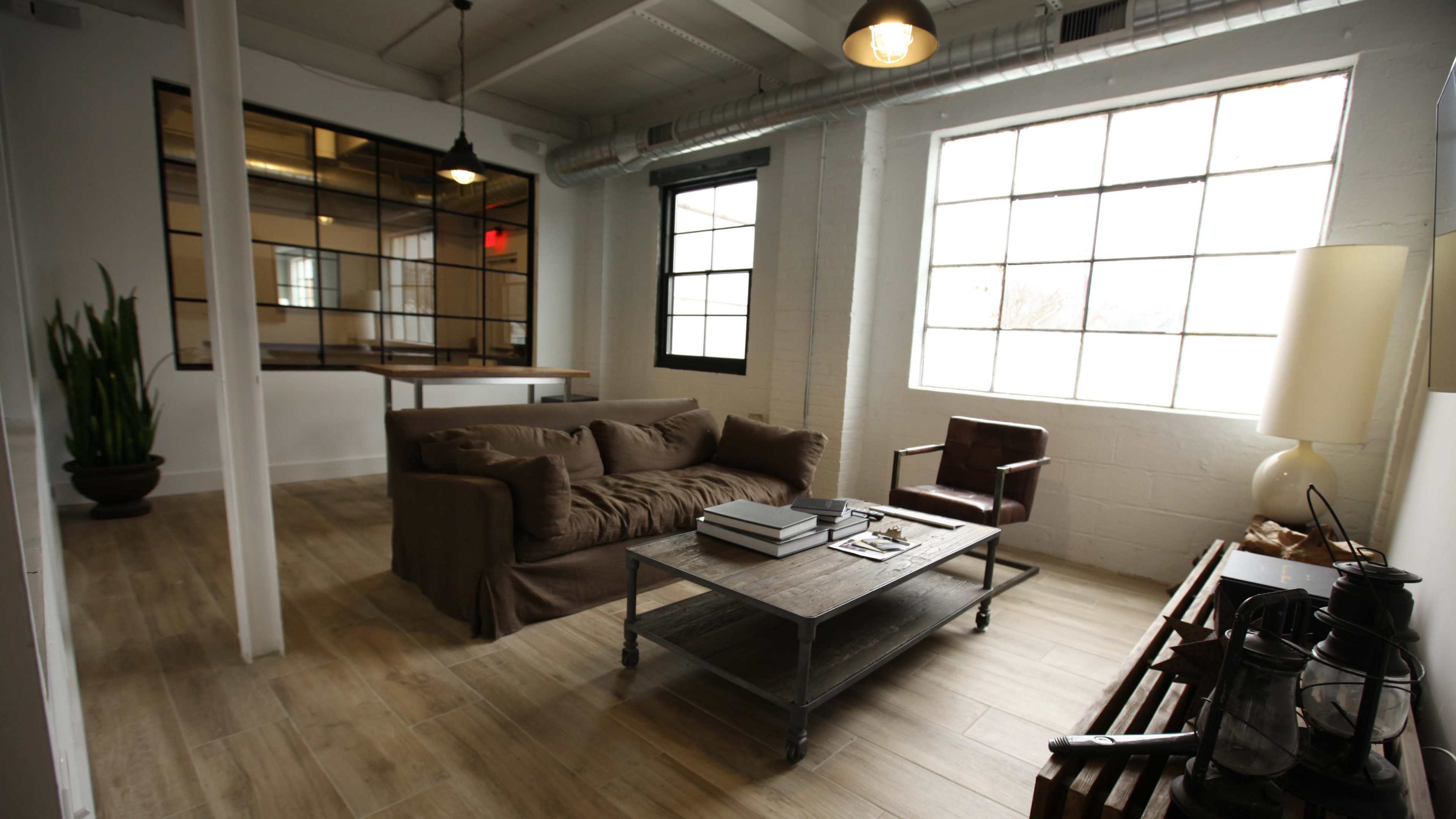 The image shows a modern living area with a brown sofa, a wooden coffee table stacked with books, an armchair, and large windows allowing natural light.