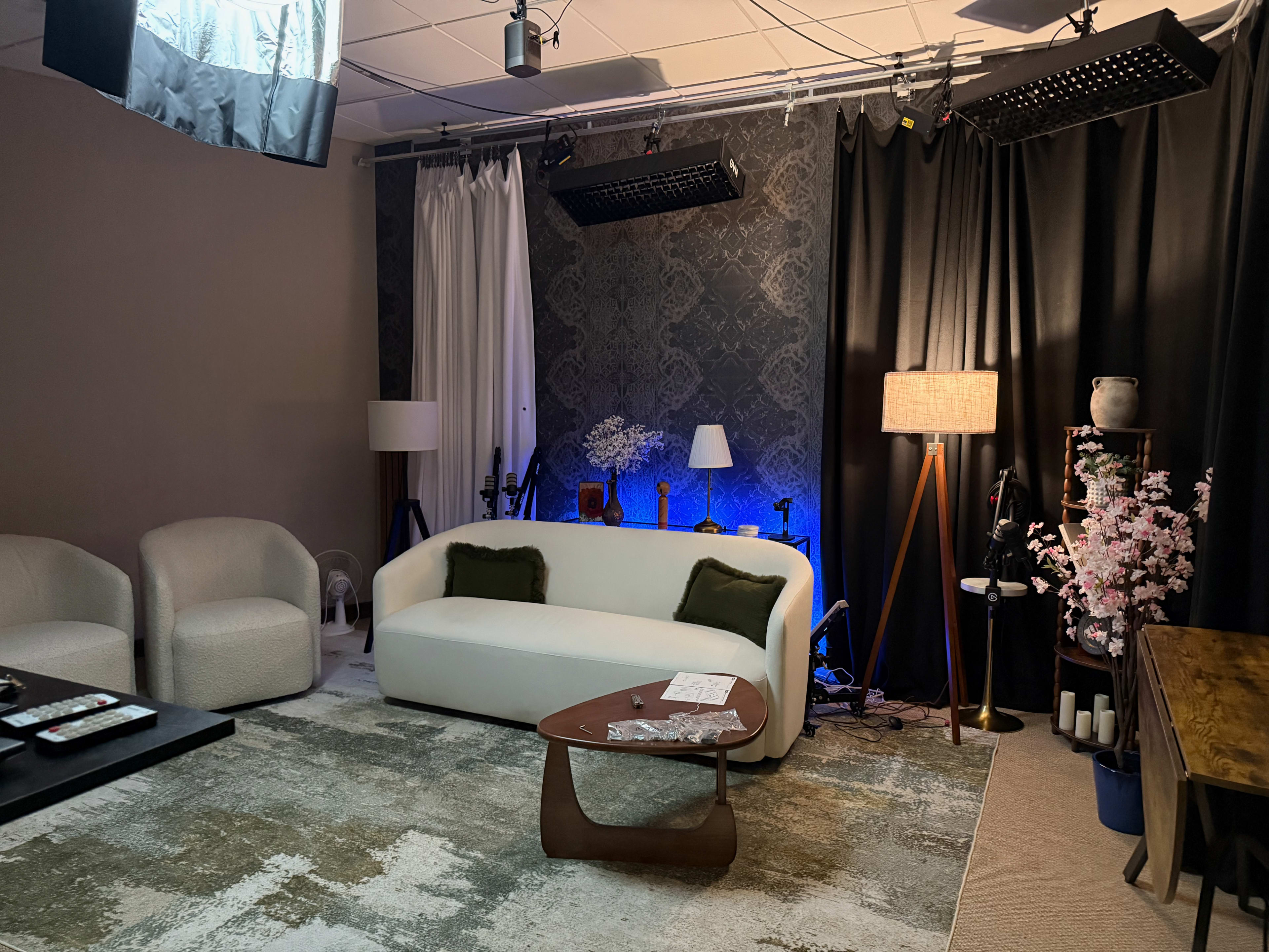 The image shows a modern studio setup featuring a white couch, a wooden coffee table, decorative lamps, and various equipment against dark curtains and a patterned wall.