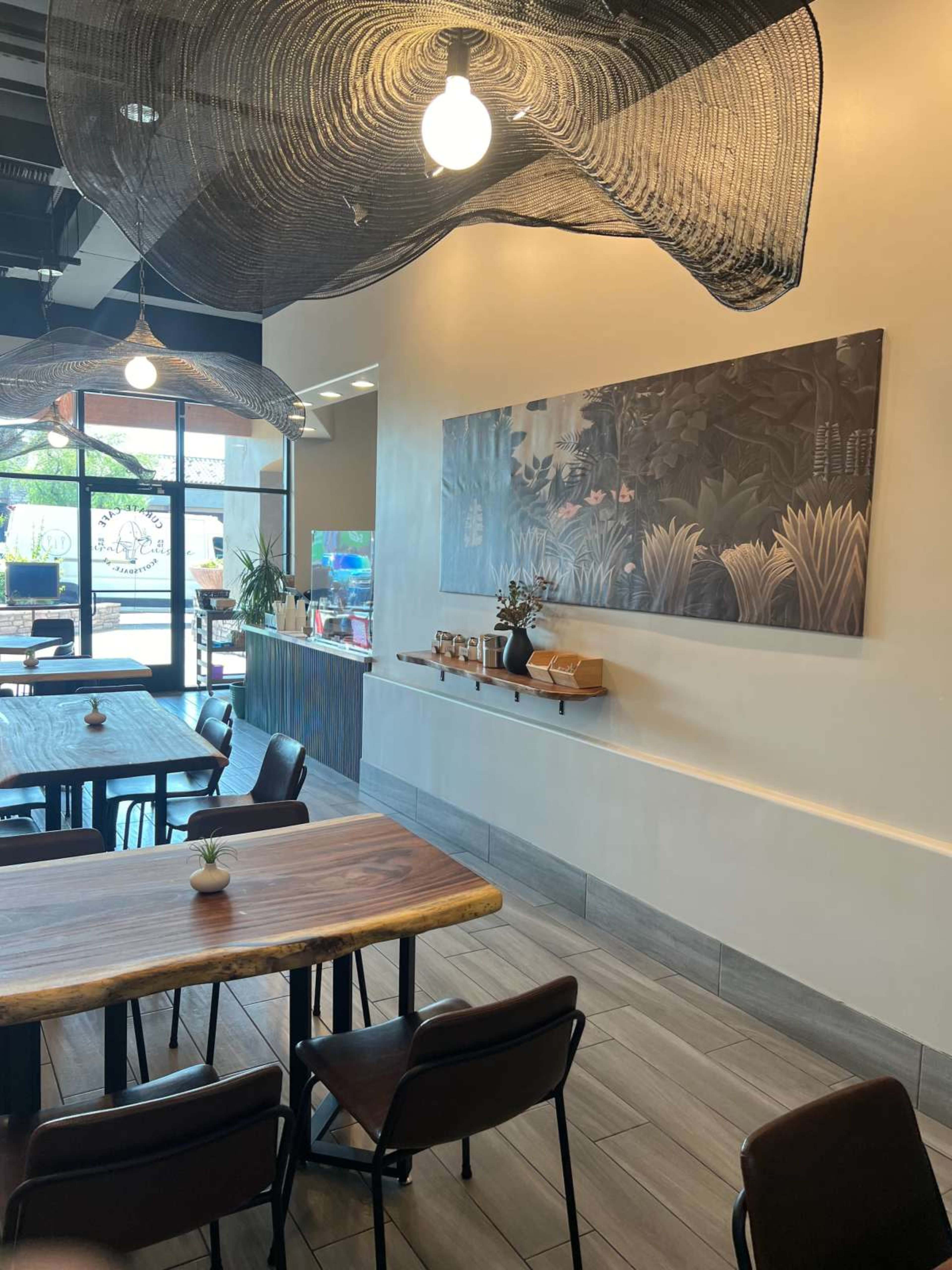 A modern café interior featuring wooden tables, black chairs, and decorative hanging elements above, with a mural on the wall and a view of the entrance.