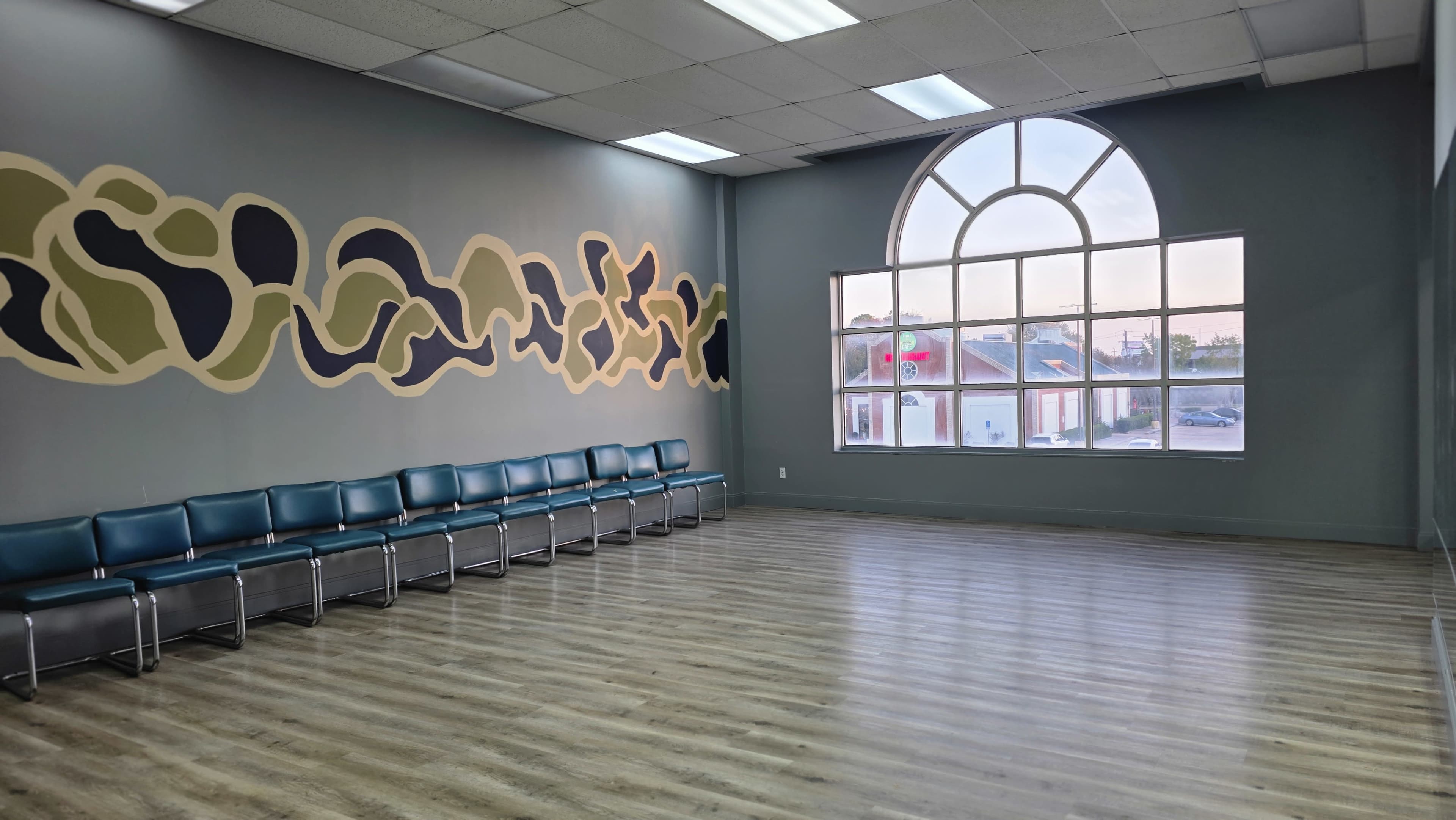 The image shows an empty waiting room with a row of blue chairs against a colorful mural on one wall and a large window letting in natural light.