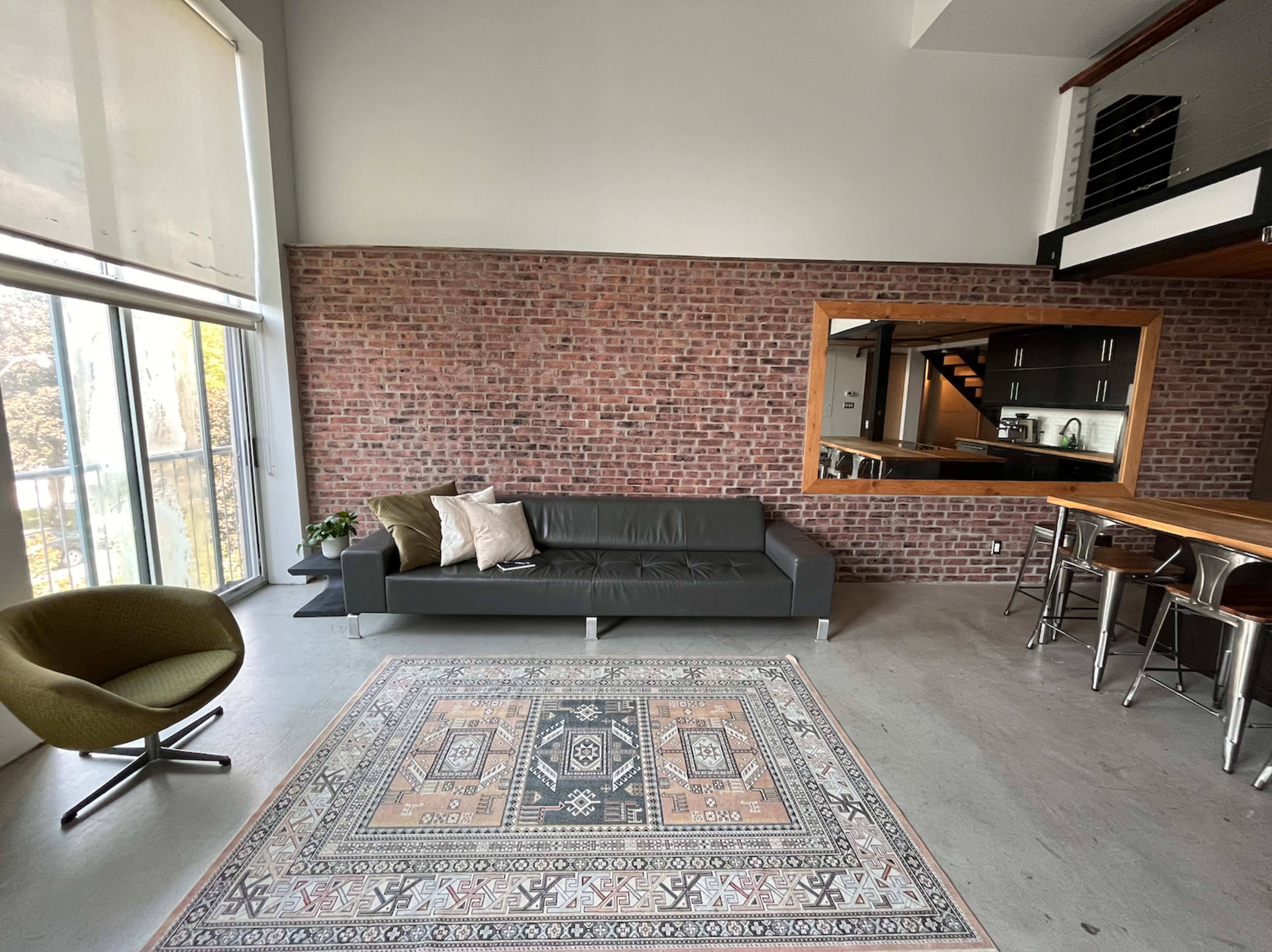 A modern living space featuring a dark sofa, a patterned area rug, a green accent chair, and exposed brick walls, with a glimpse of a kitchen area in the background.