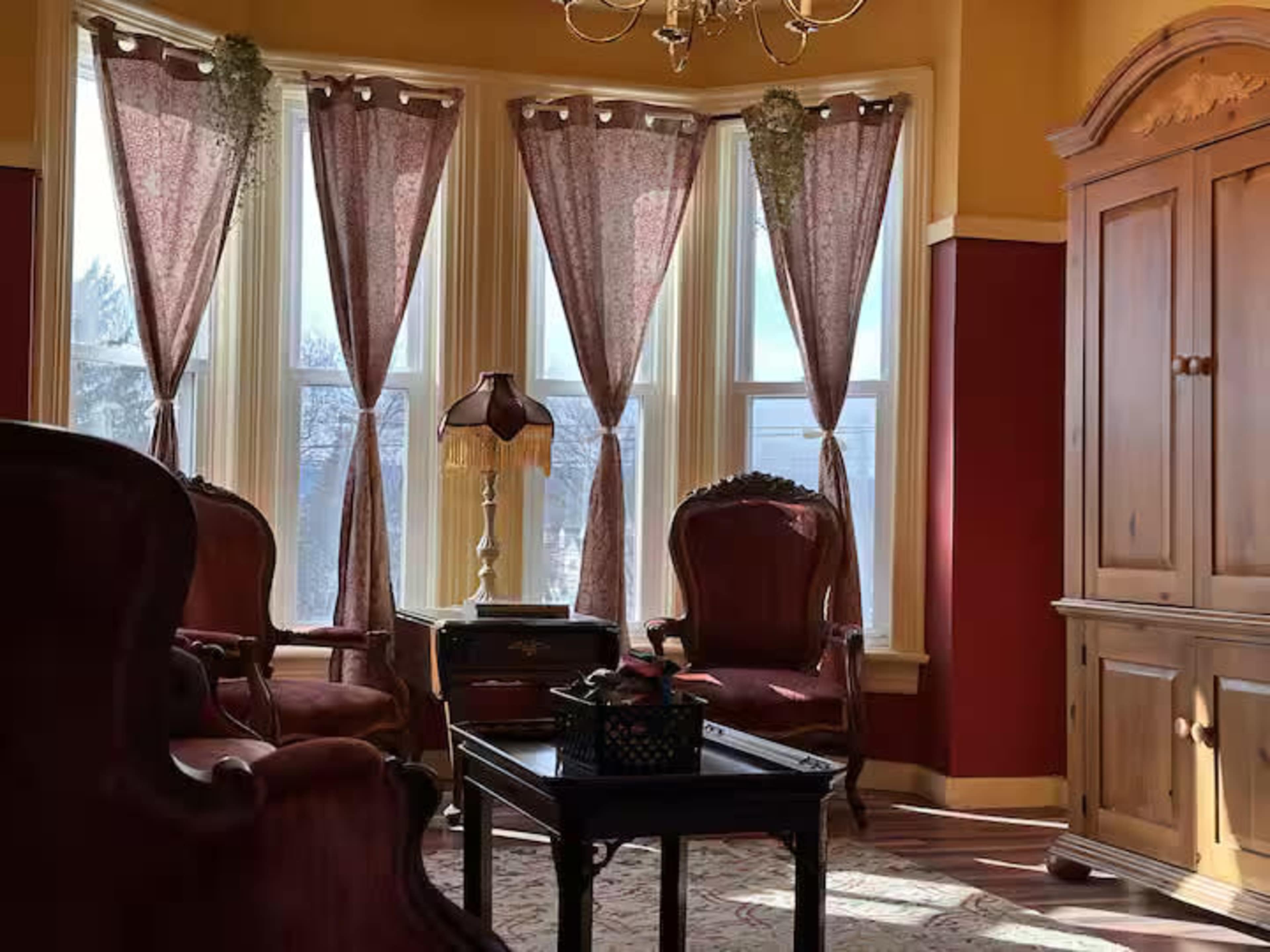 The room features a pair of vintage upholstered chairs, a wooden cabinet, and large windows adorned with draped curtains, allowing natural light to fill the space.