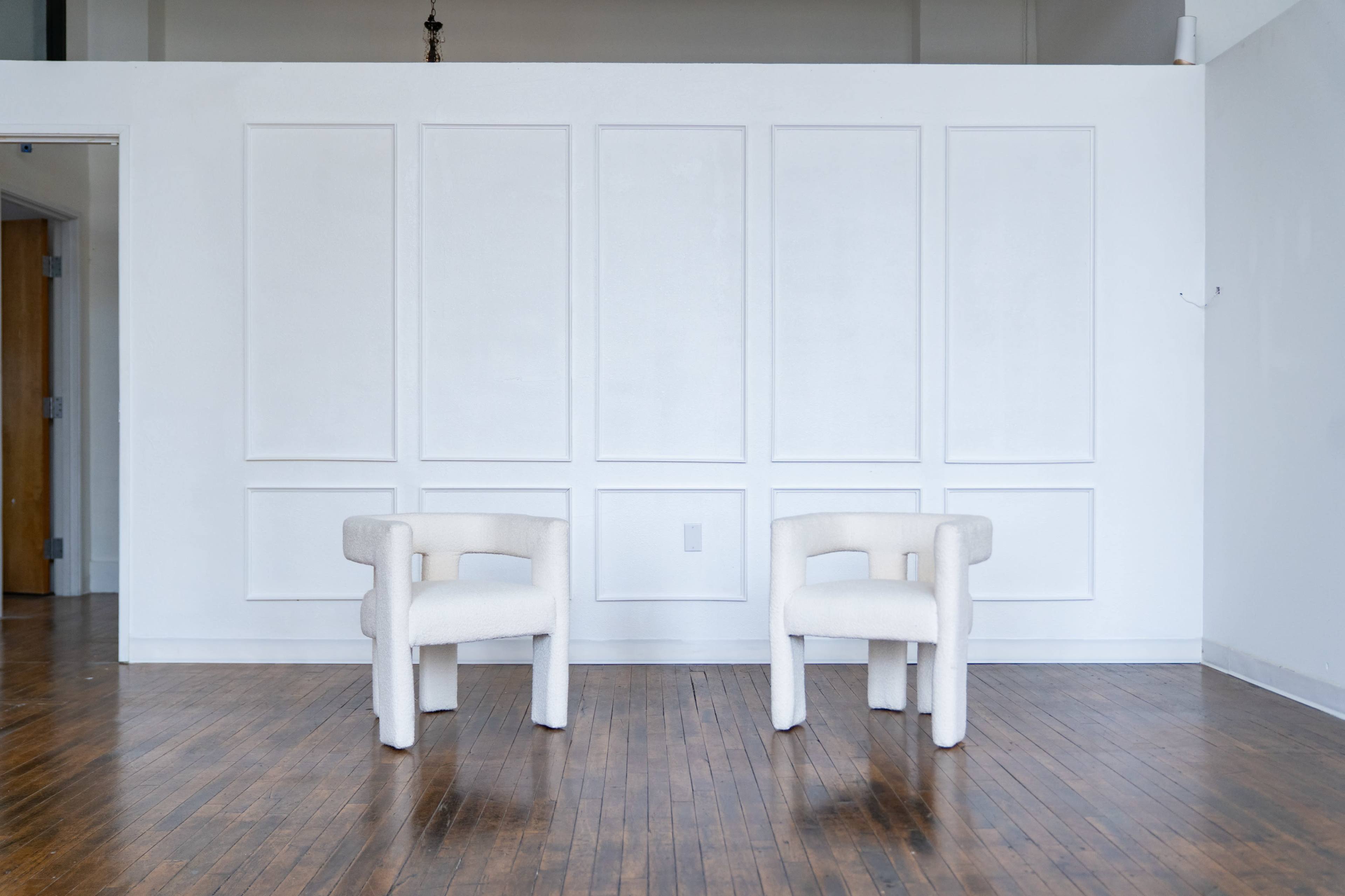 Two white, upholstered armchairs are positioned in front of a plain, white-paneled wall in a spacious room with wooden flooring.