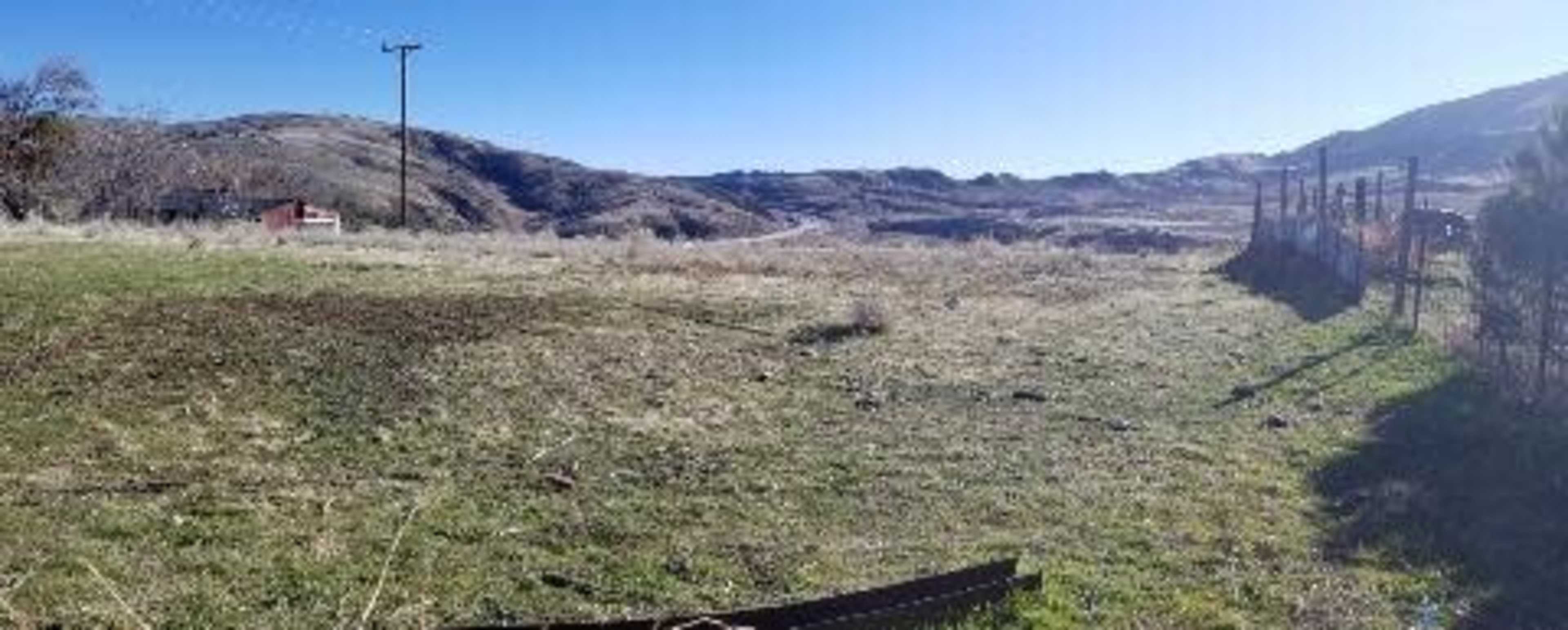 A grassy field bordered by a fence, with distant hills and a road visible in the background.