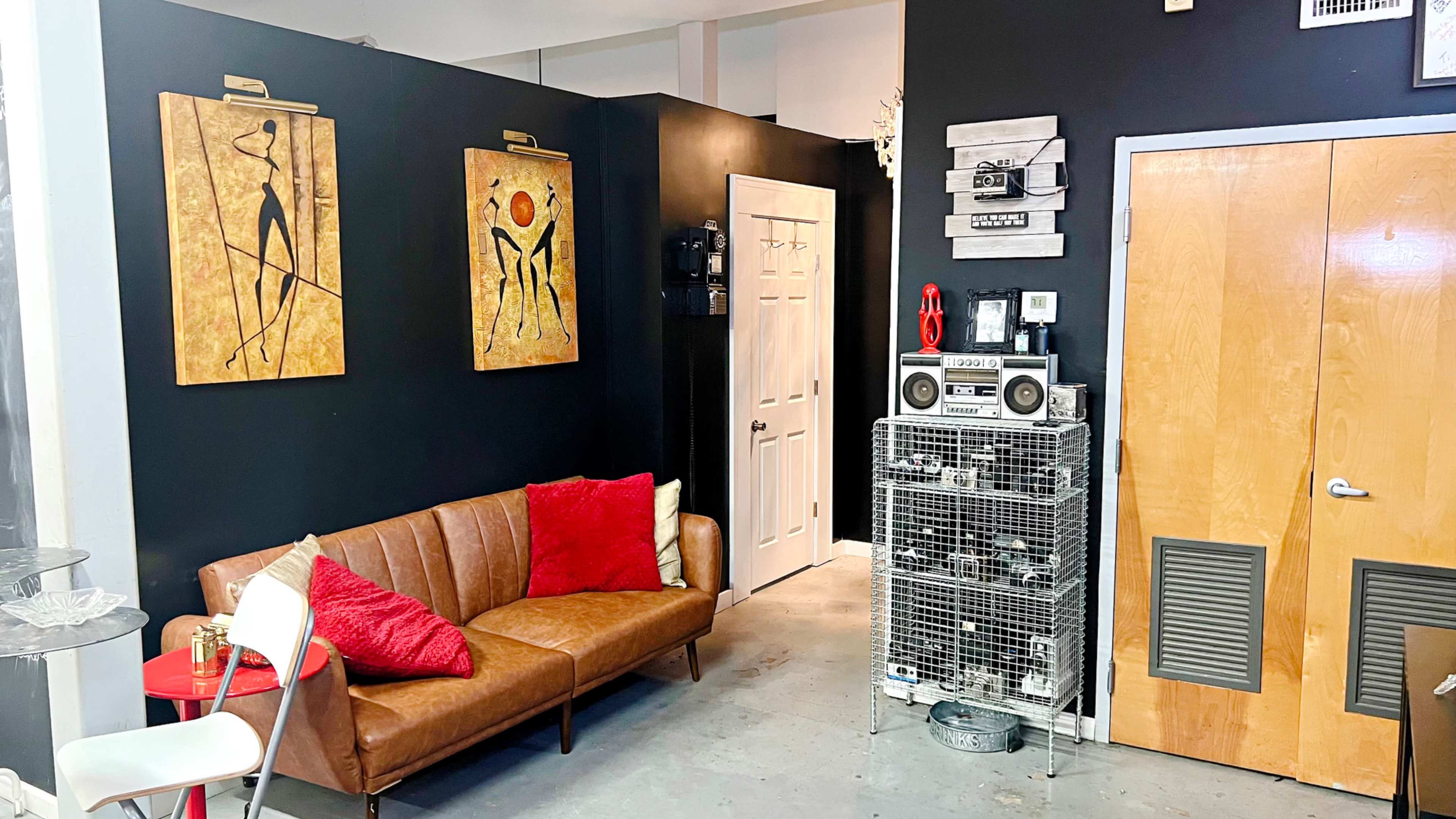 The image shows a modern interior space featuring a brown leather sofa with red cushions, two abstract wall paintings, and a vintage audio system next to a wooden door.
