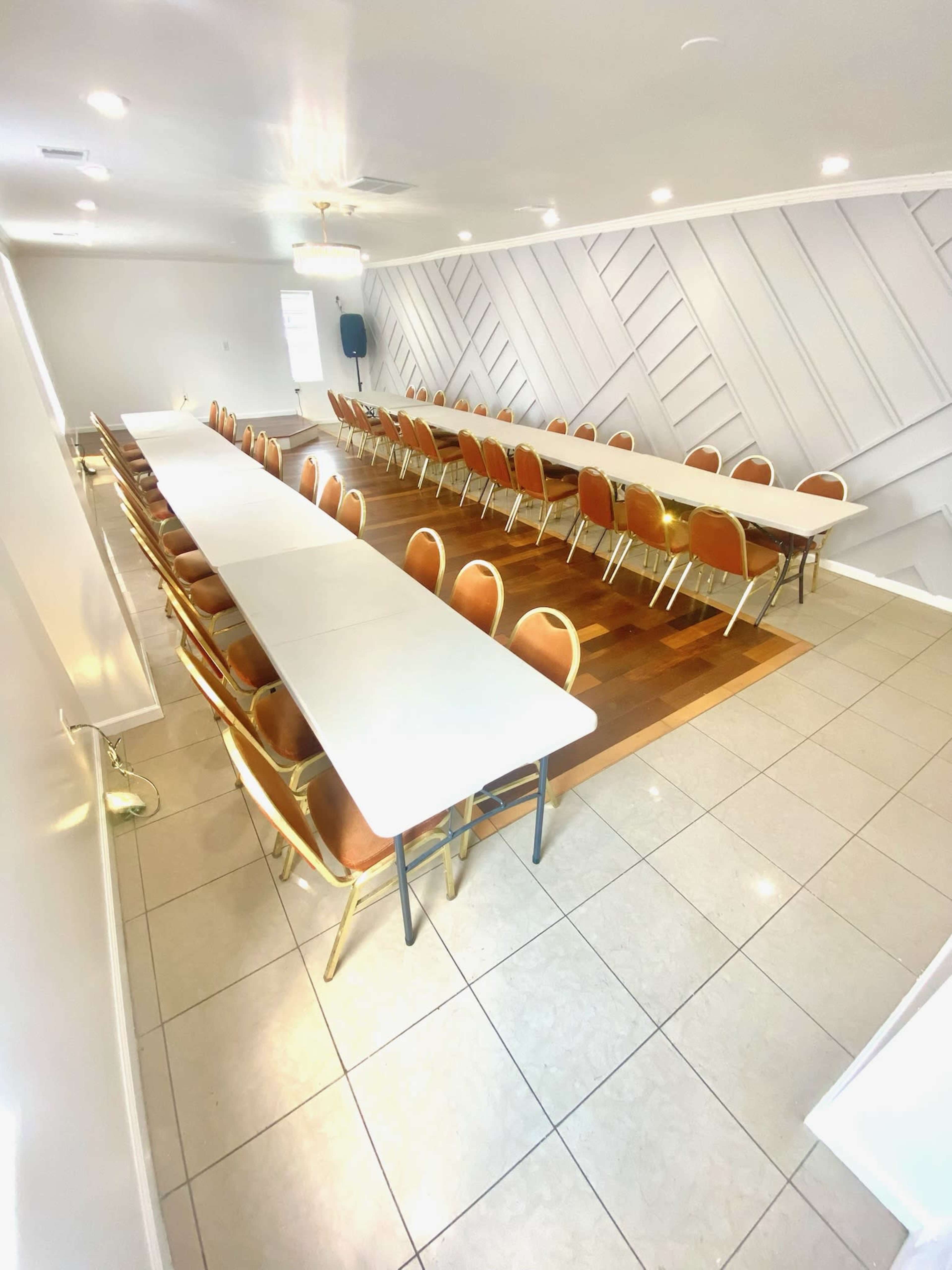 The image shows a large meeting room arranged with multiple long tables and chairs in rows, set on a tile and wooden floor.