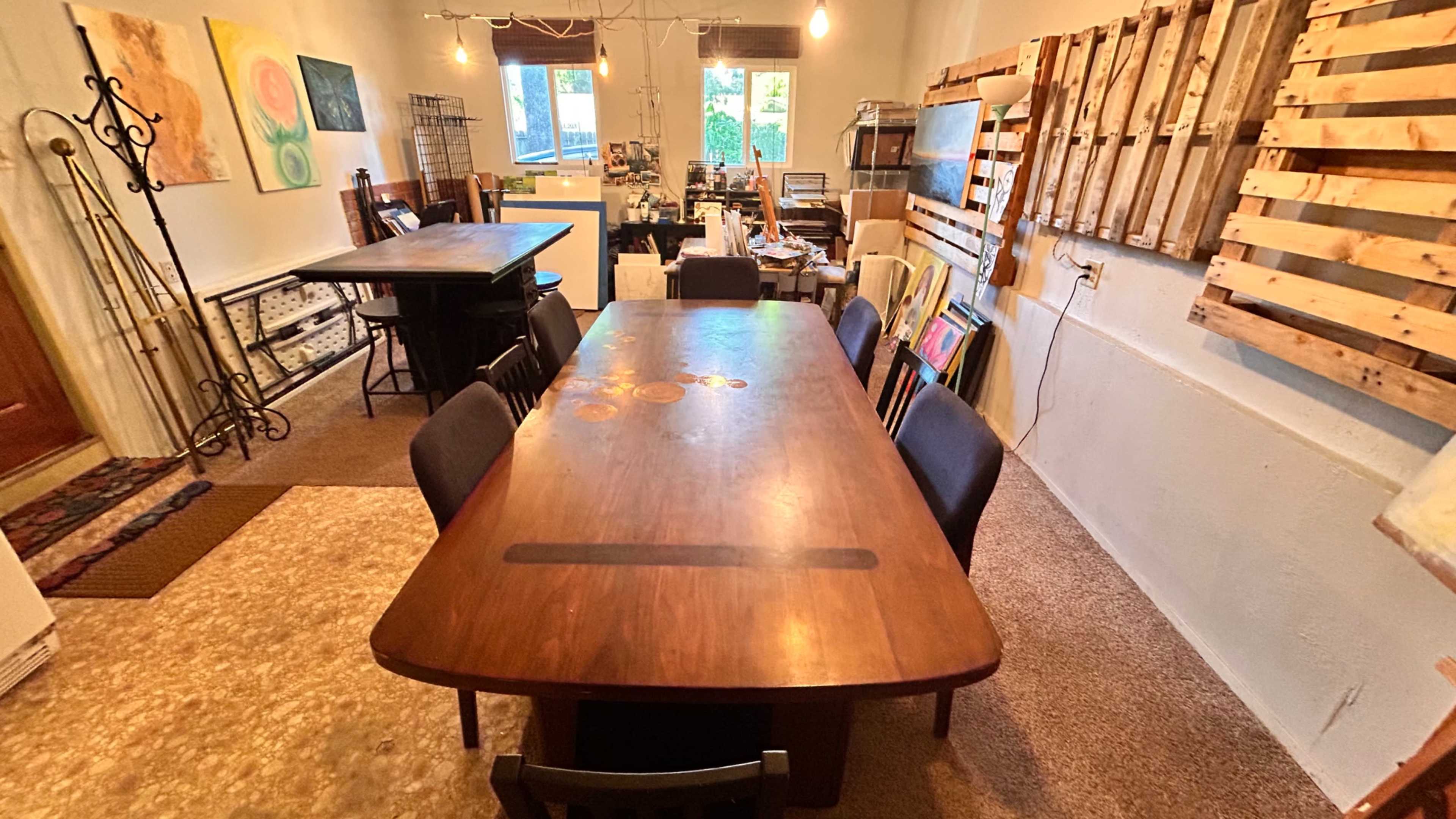 The image features a spacious room with a large wooden dining table surrounded by black chairs, along with various artworks and wooden pallets mounted on the walls.