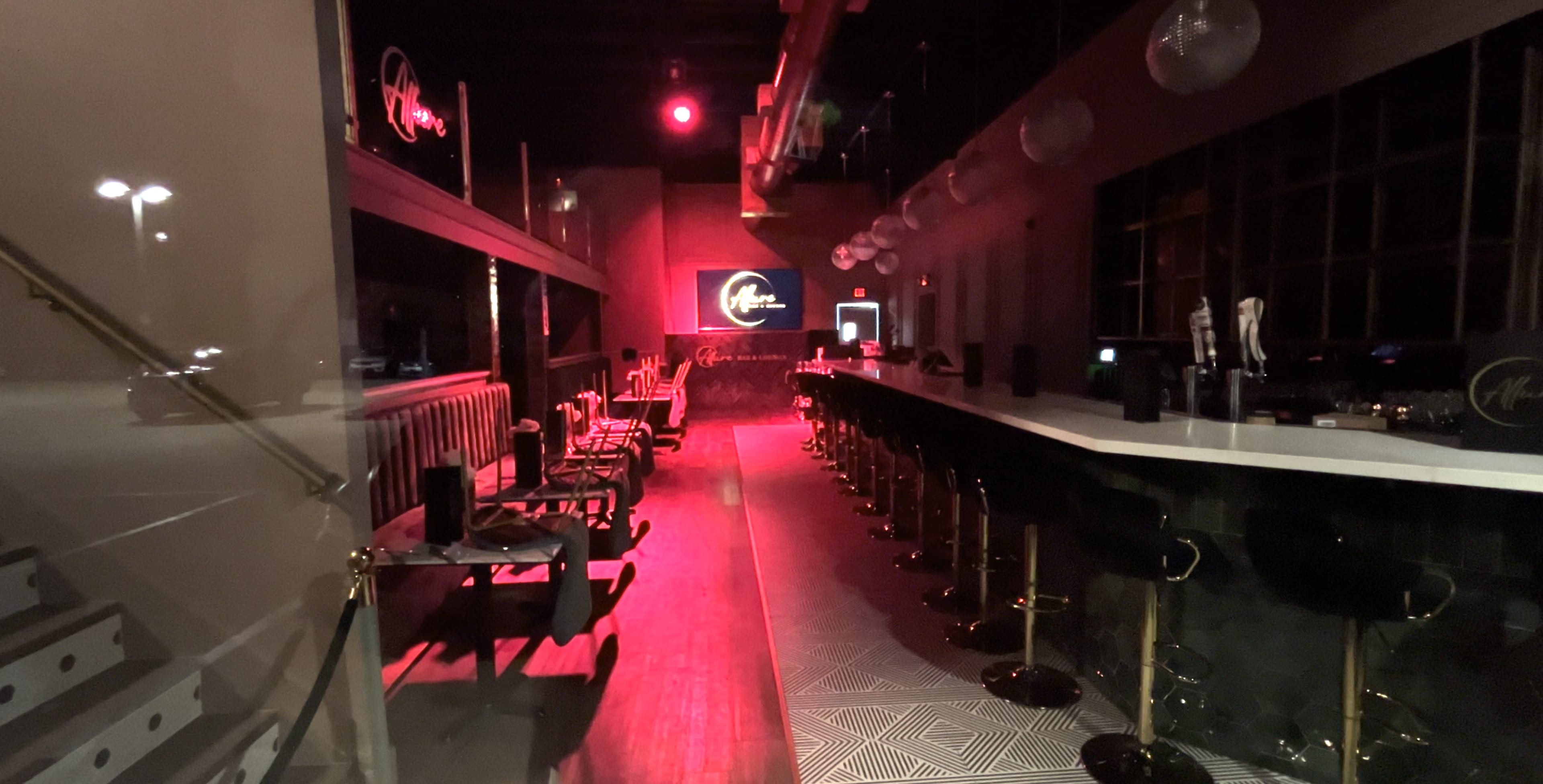 The image shows a dimly lit bar with a long counter and black chairs, illuminated by red and white lights.
