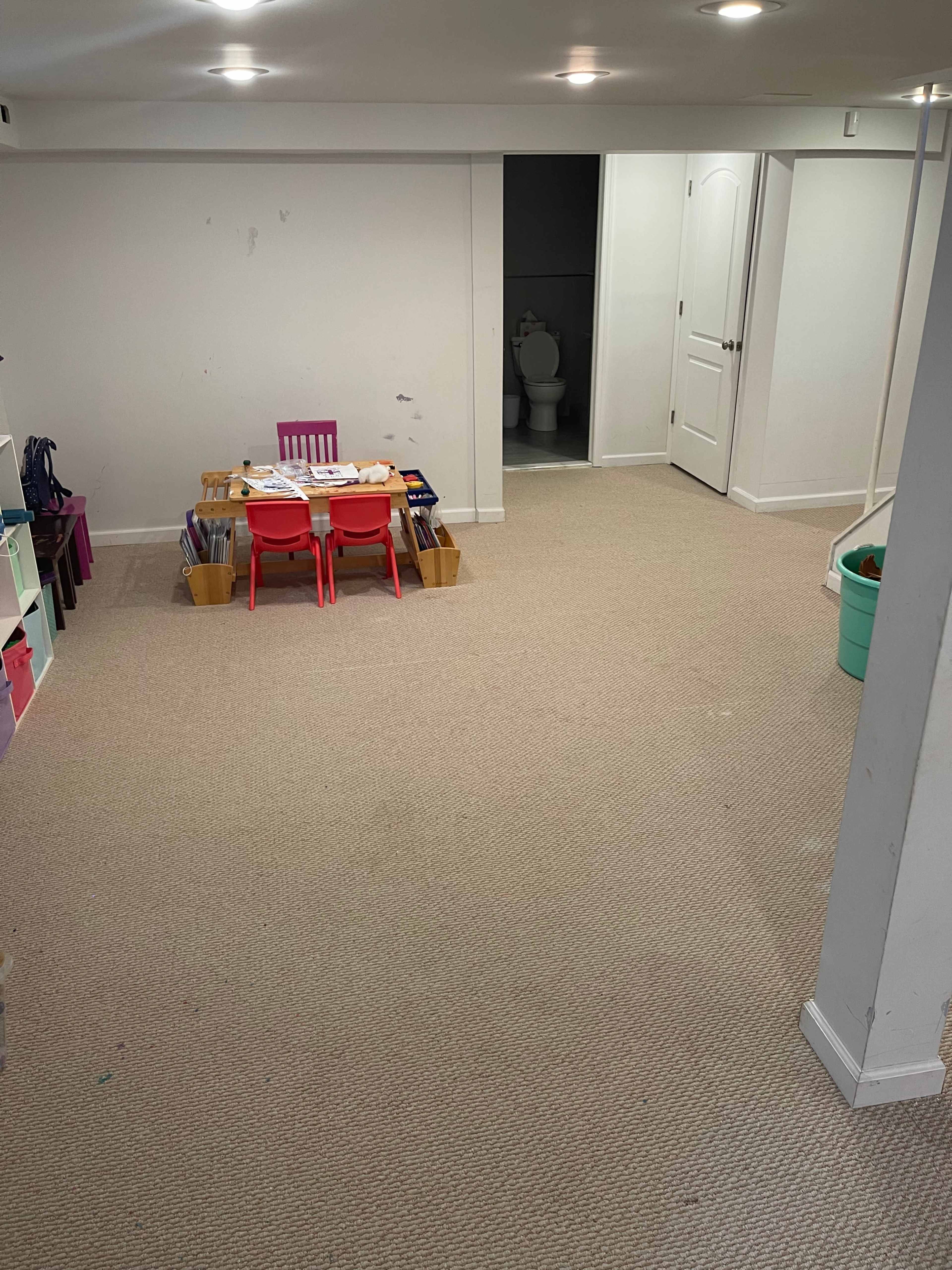 A spacious, carpeted basement features a small table with chairs surrounded by storage bins and a bathroom door in the background.