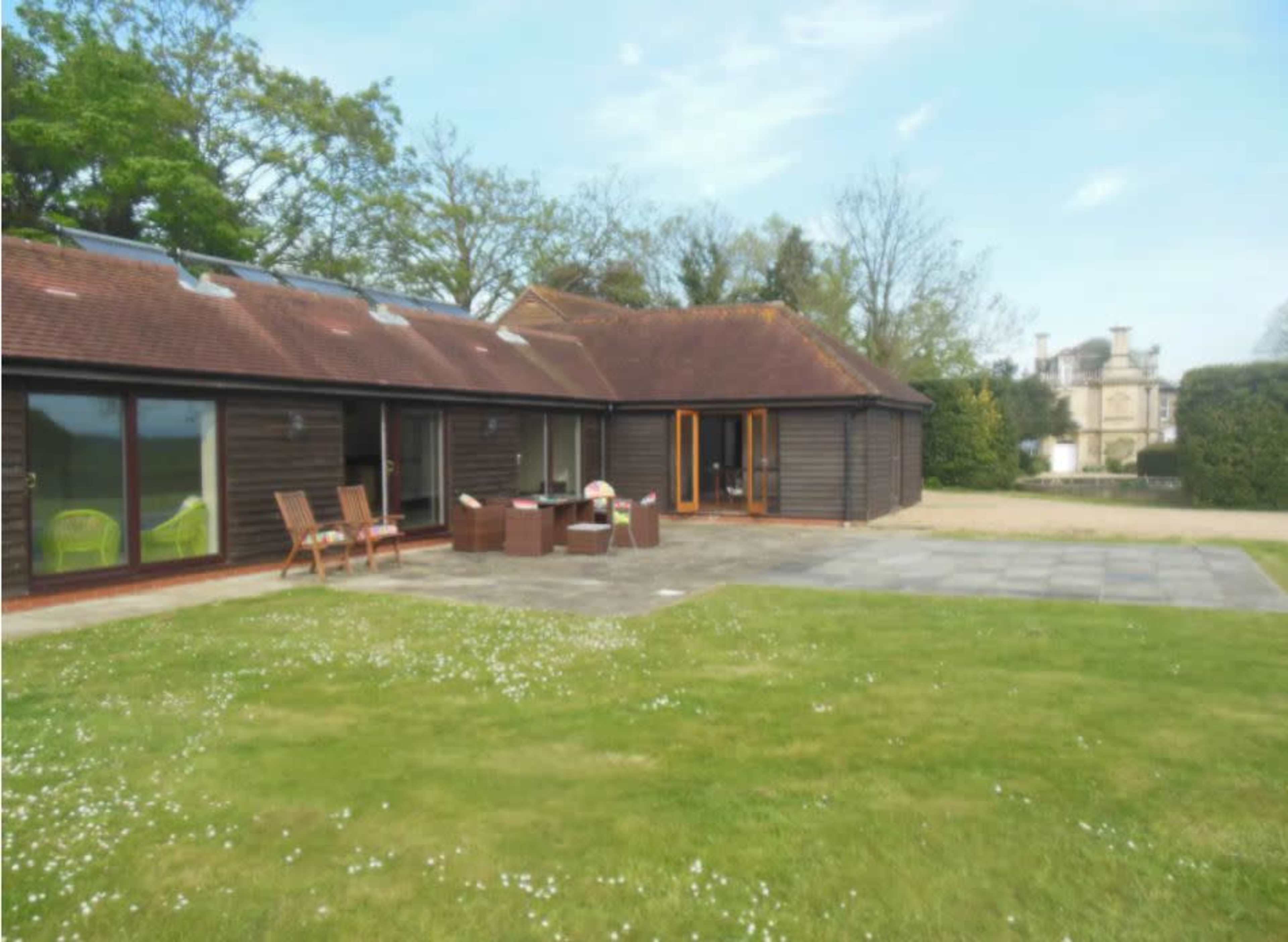 A modern single-story wooden house features large glass windows and a stone patio overlooking a manicured lawn.