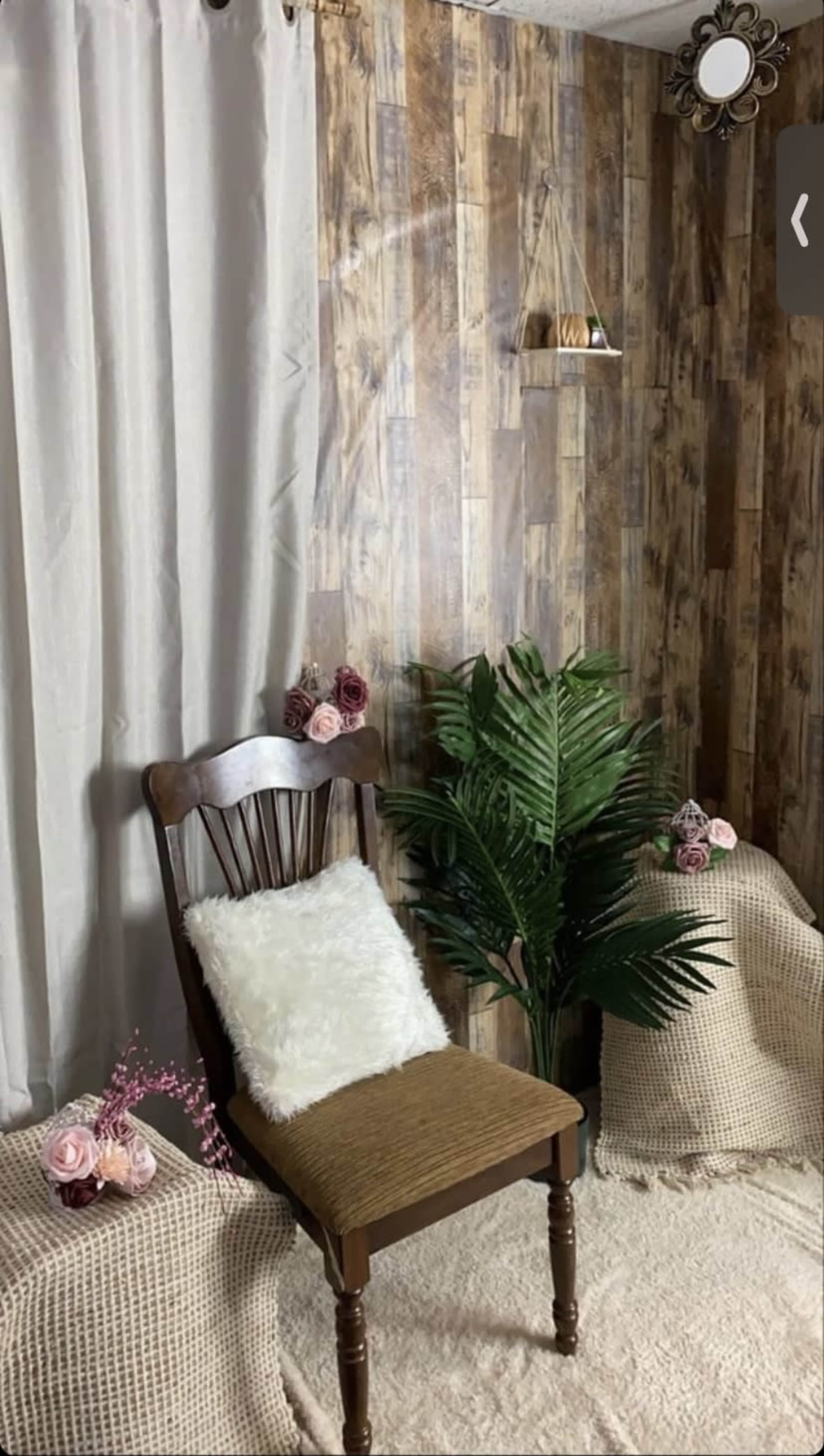 A cozy room corner with a wooden chair, a soft pillow, potted plants, and decorative flowers against a rustic wood-paneled wall.