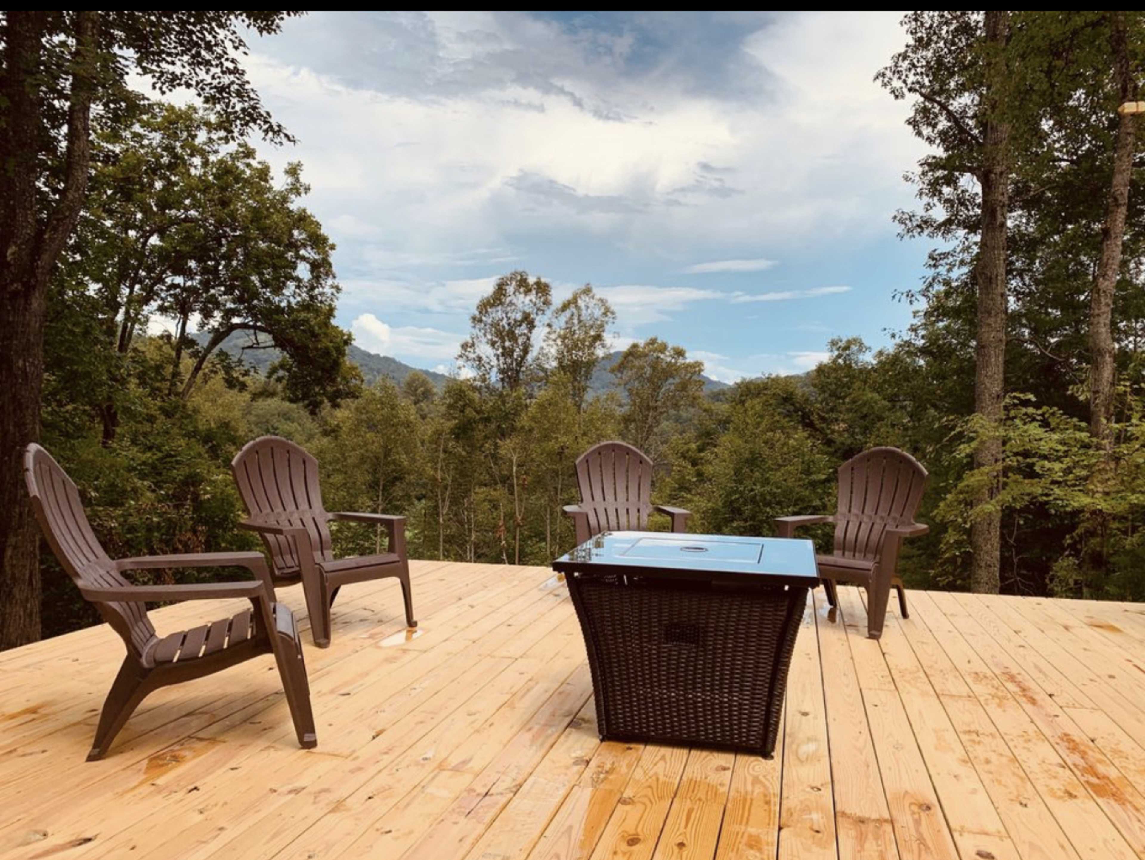 A wooden deck overlooks a forested landscape, featuring four brown chairs arranged around a blue coffee table.