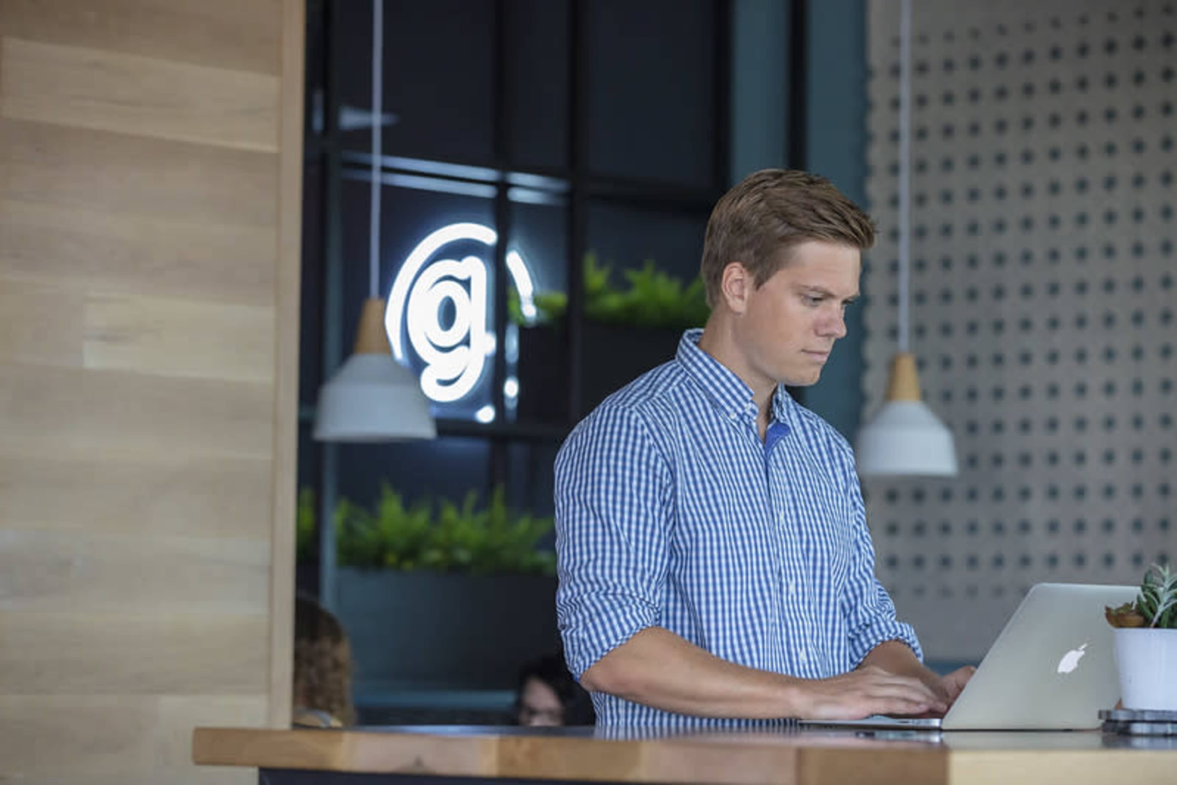 A man works on a laptop at a modern workspace with a bright sign in the background.