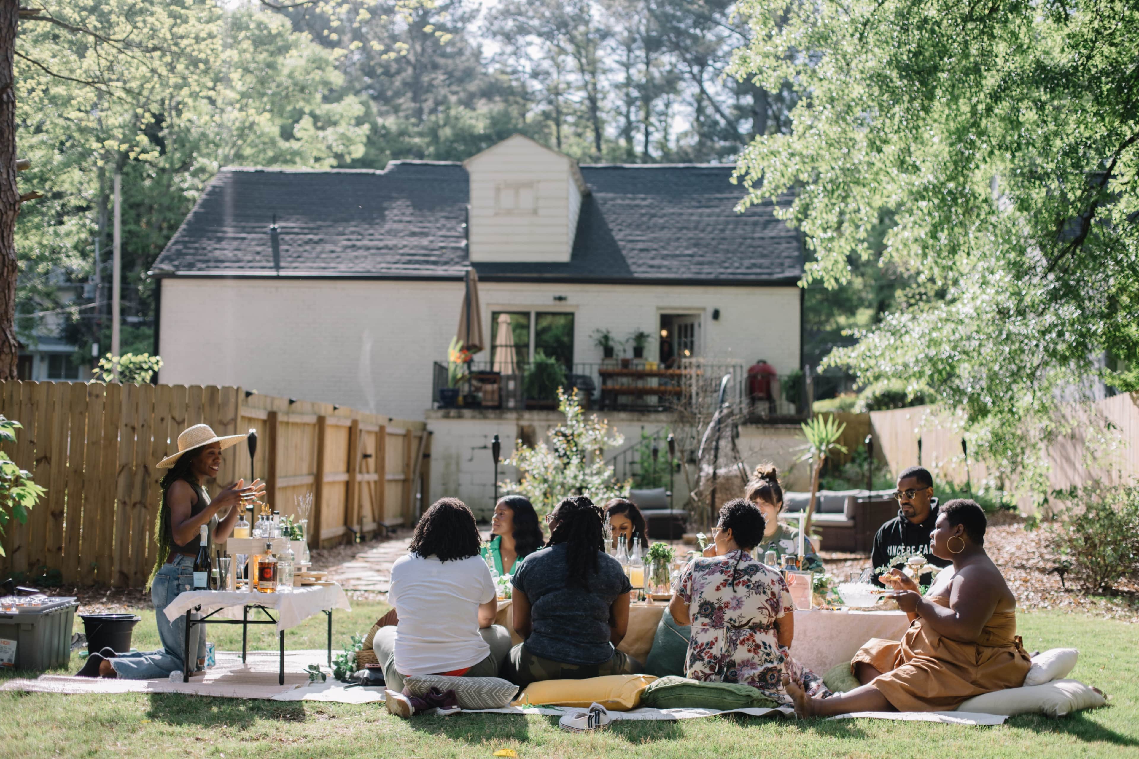 A group of people gather around a table in a garden, enjoying food and drinks on a sunny day.