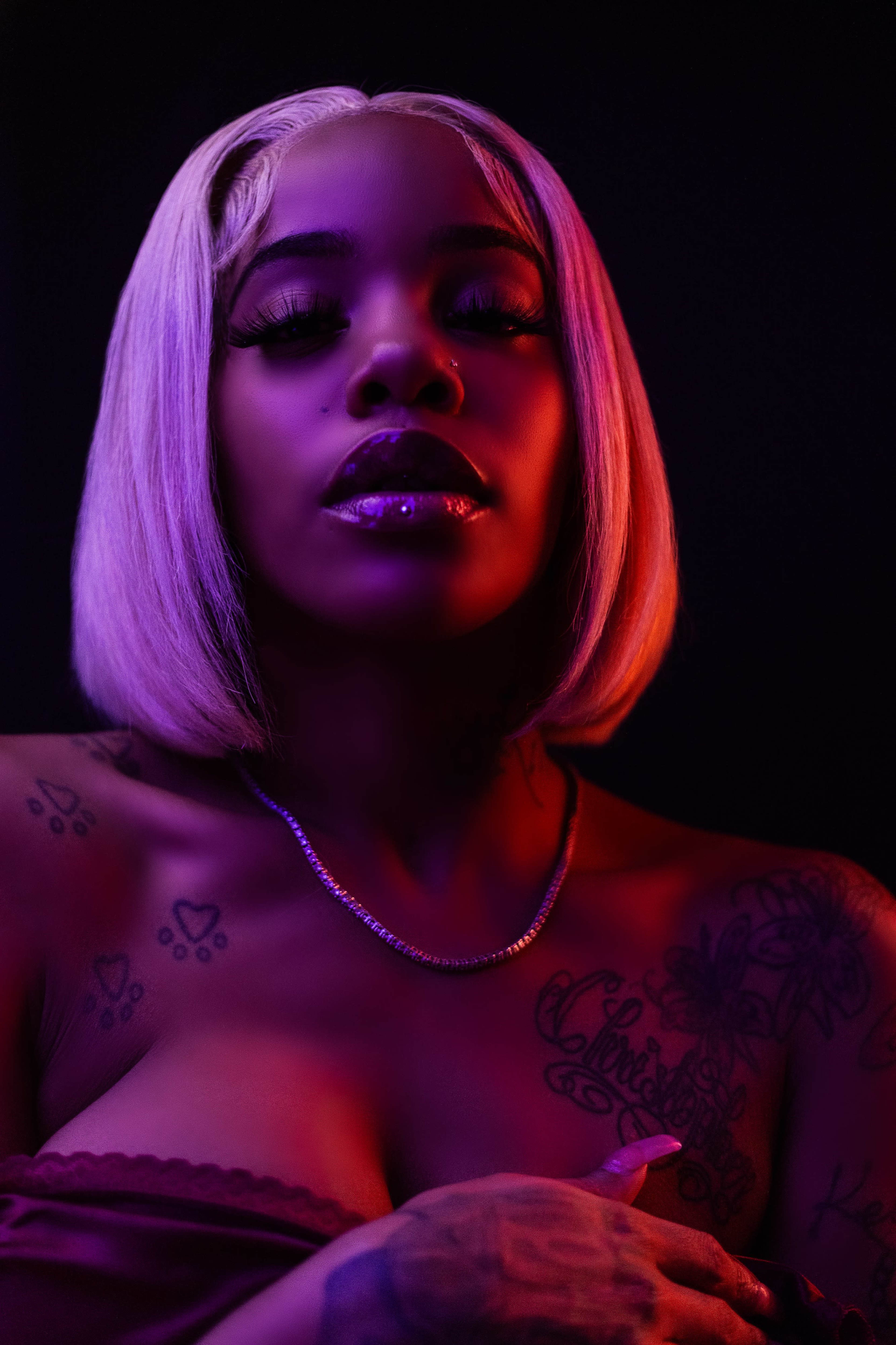 A woman with a short blonde bob hairstyle poses in front of a dark background, adorned with tattoos and wearing a low-cut outfit.