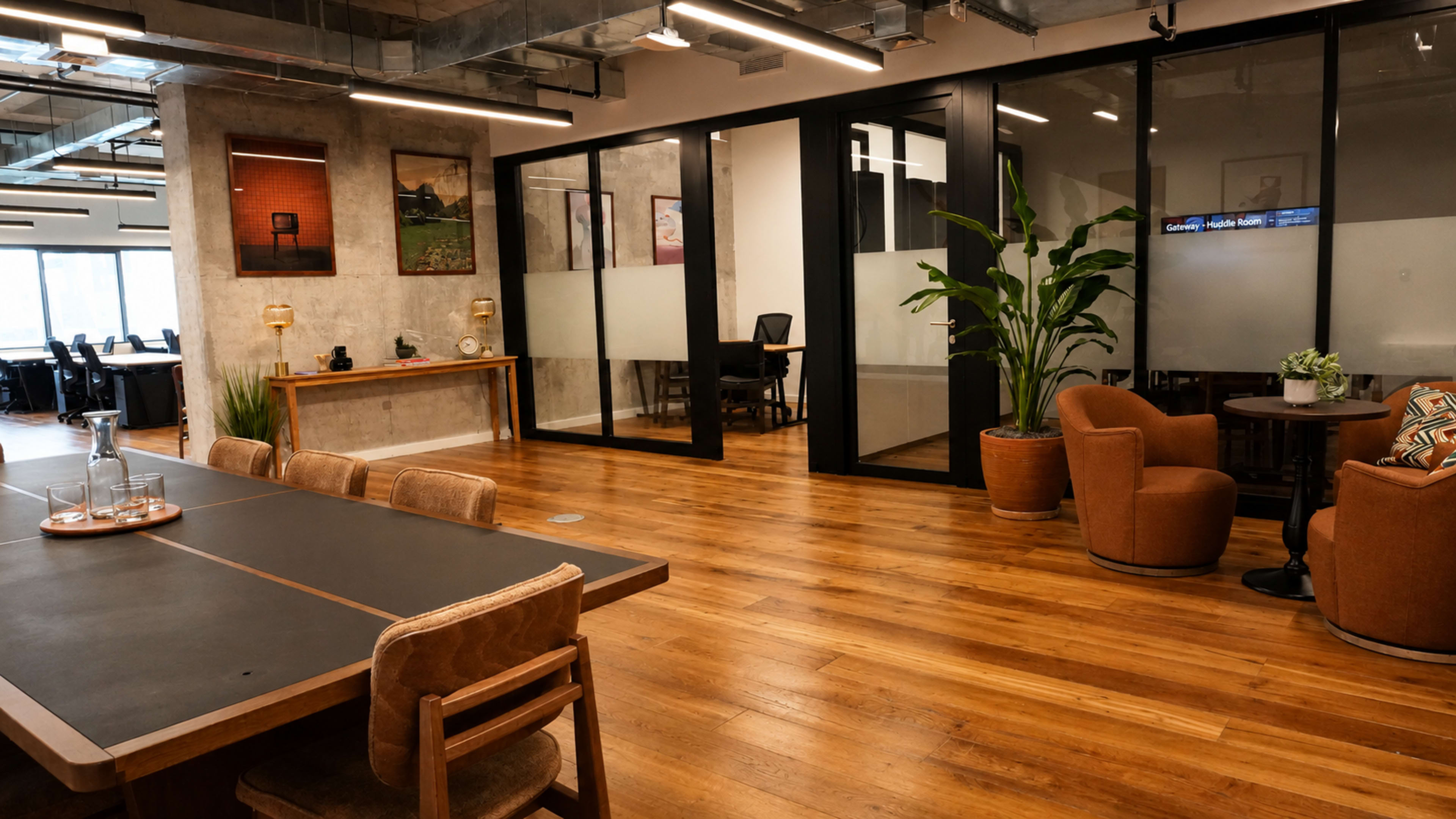 The image shows a modern office space with a wooden floor, featuring a communal table, cozy seating areas, and glass-partitioned meeting rooms.