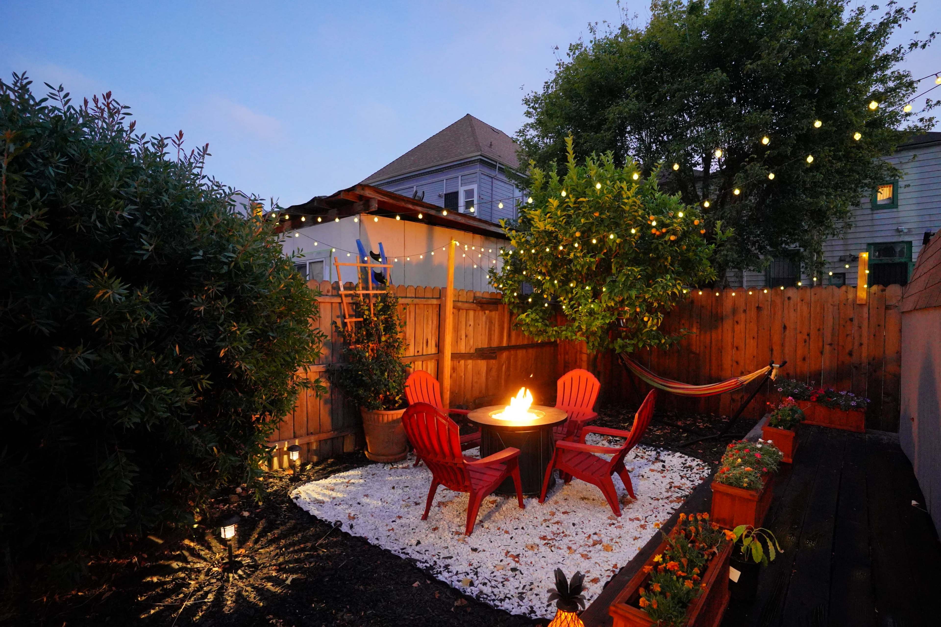 A small outdoor patio features red chairs around a fire pit, with potted plants along the edges and string lights overhead.