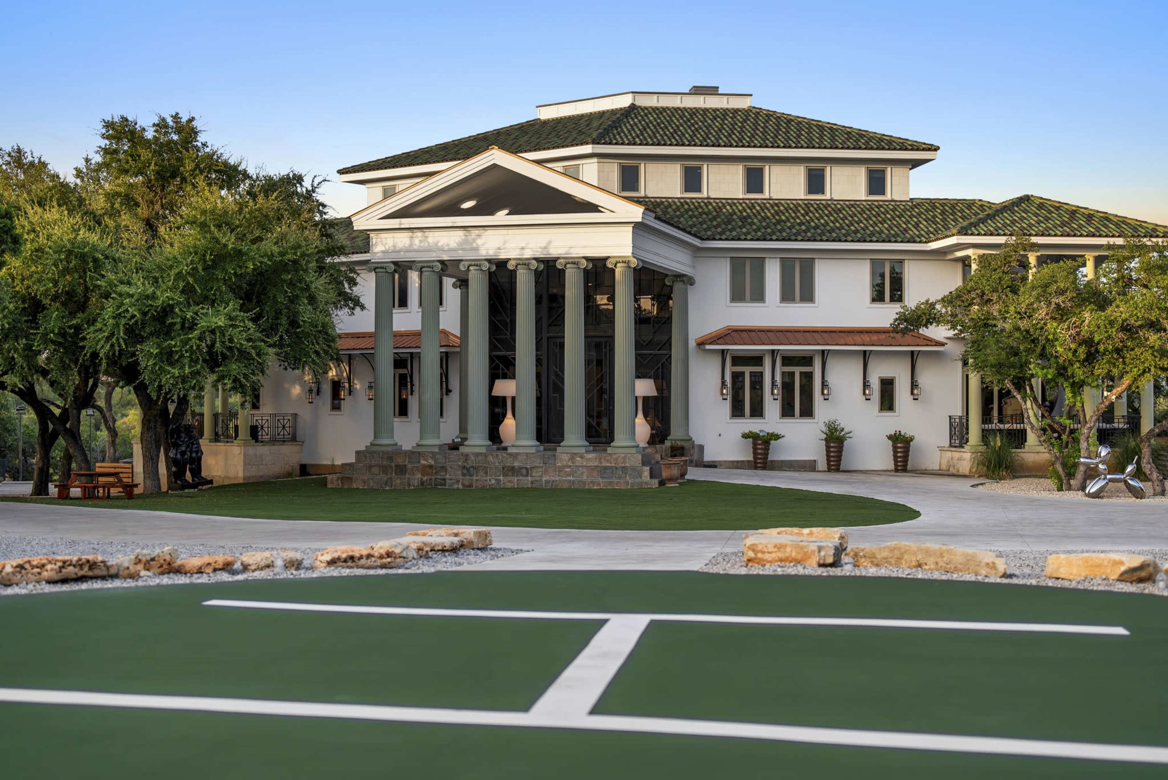 A large, modern mansion featuring a series of columns at the entrance and manicured landscaping.