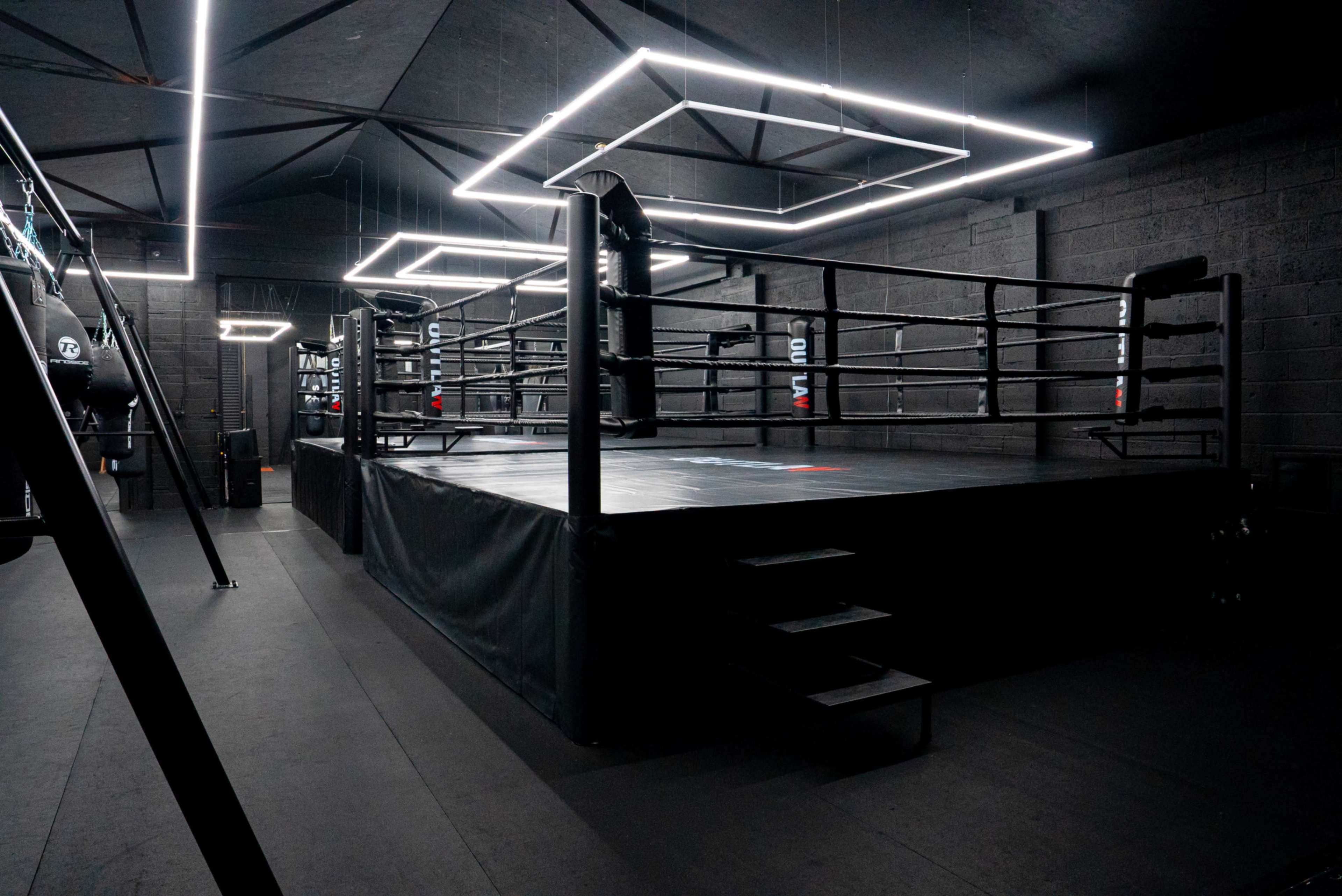 The image shows a boxing ring in a dimly lit gym, illuminated by modern rectangular light fixtures hanging from the ceiling.