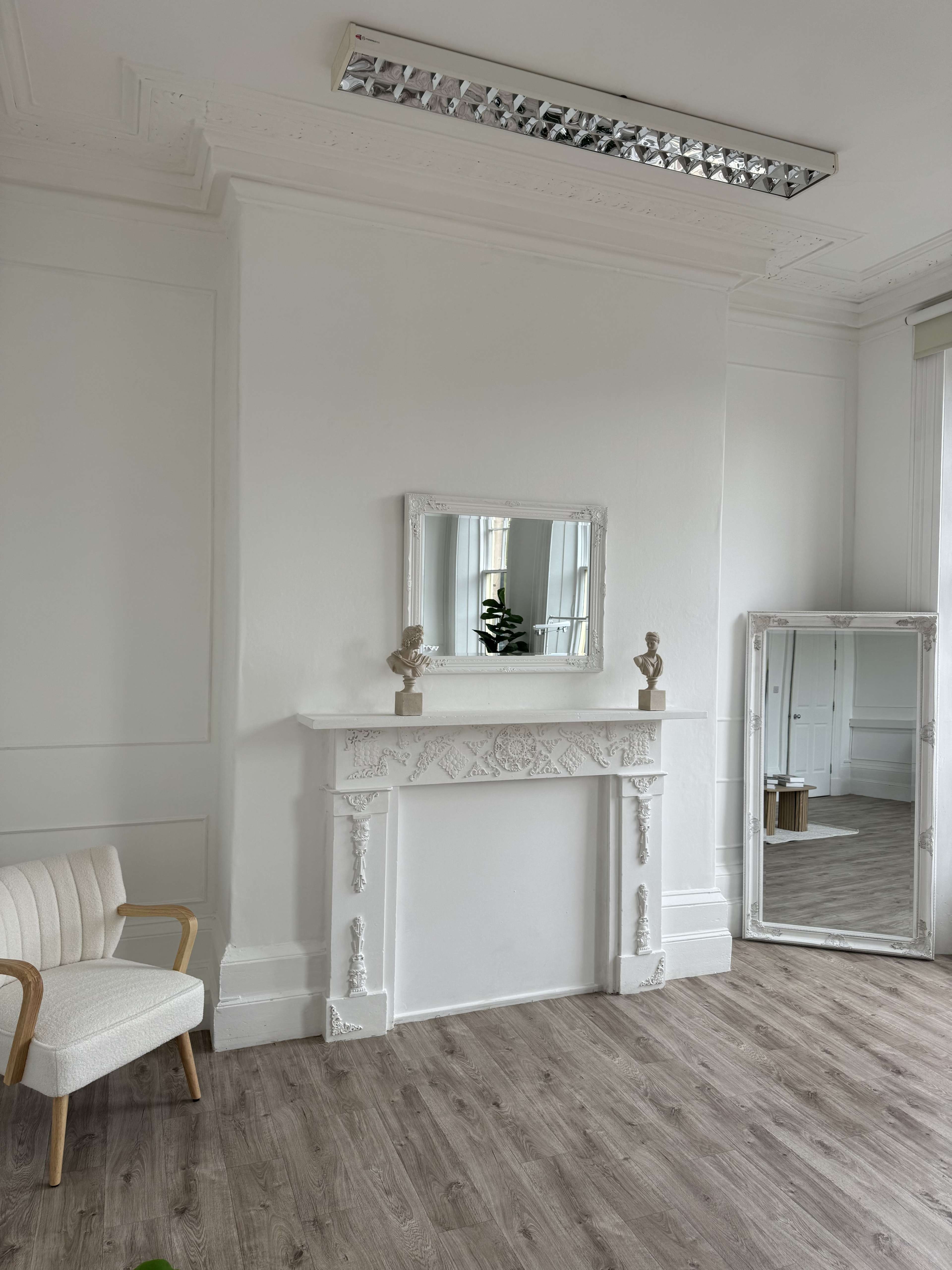 The image shows a bright and minimalist room featuring a white fireplace with decorative details, a mirror above it, a chair in the corner, and a large mirror along the opposite wall.