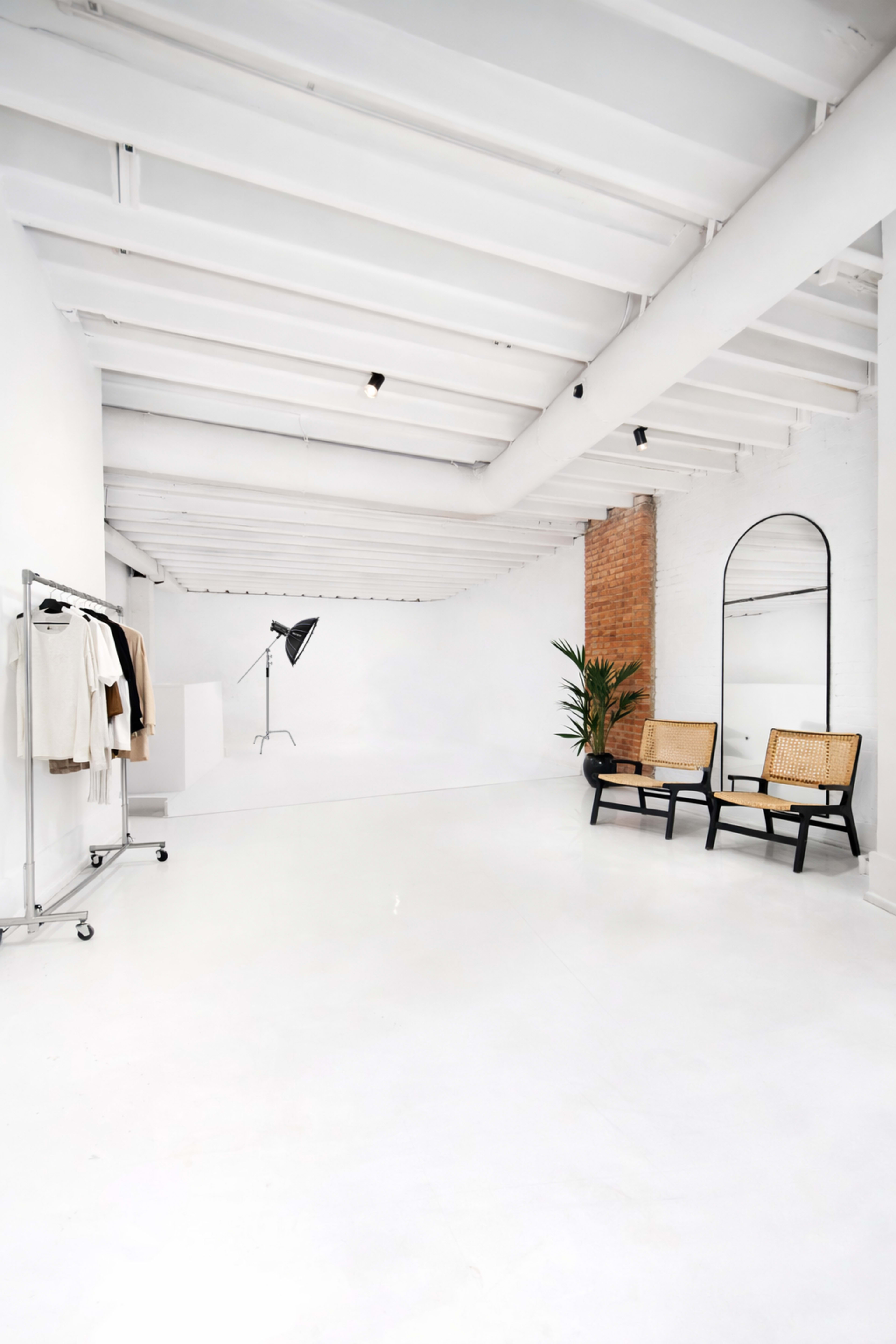 The image shows a minimalist studio space with white walls and flooring, featuring two rattan chairs, a tall mirror, a floor lamp, and a clothing rack with garments.