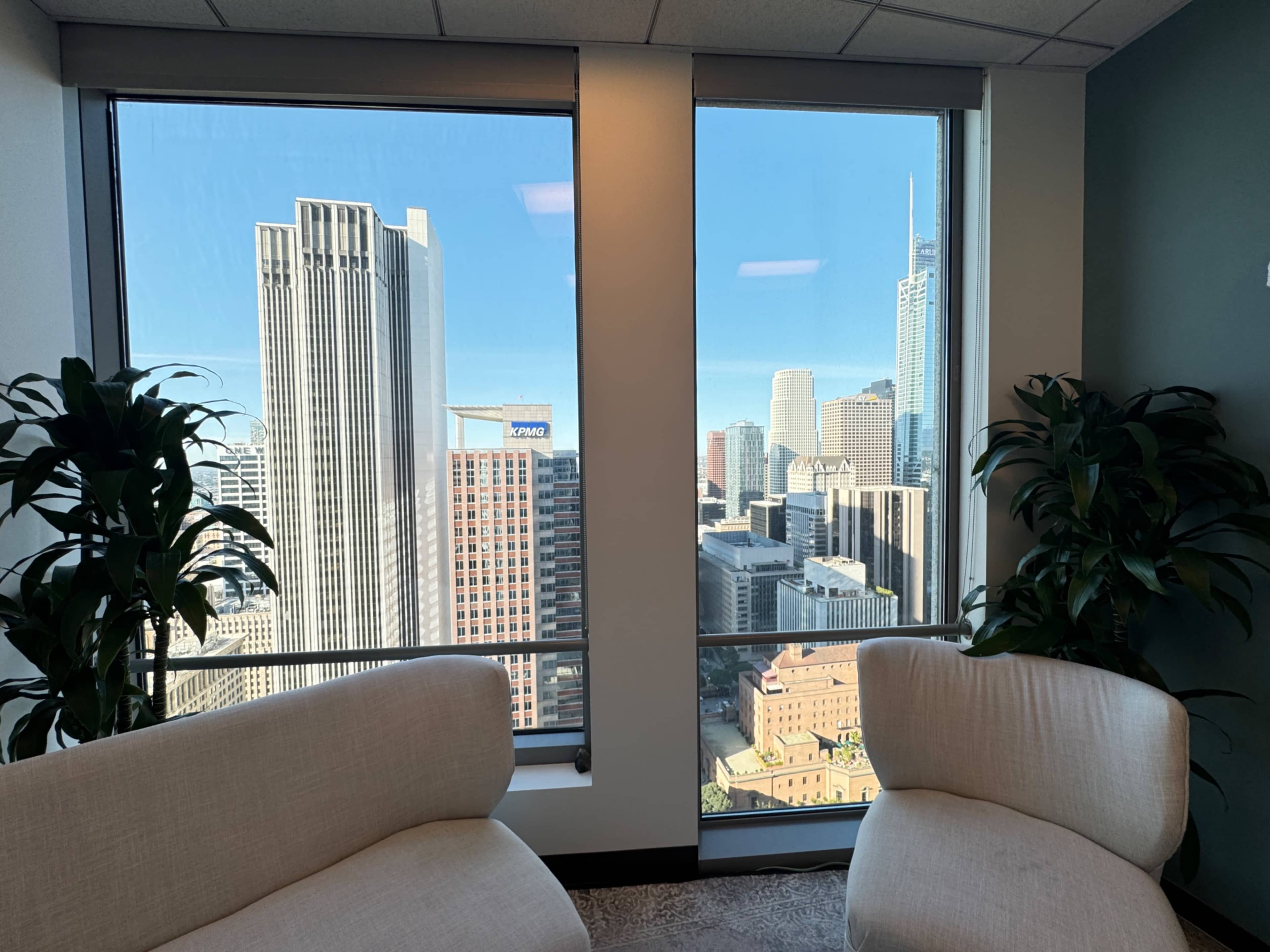 The image shows a modern office with two light-colored chairs positioned near large windows that offer a view of a city skyline featuring tall buildings.