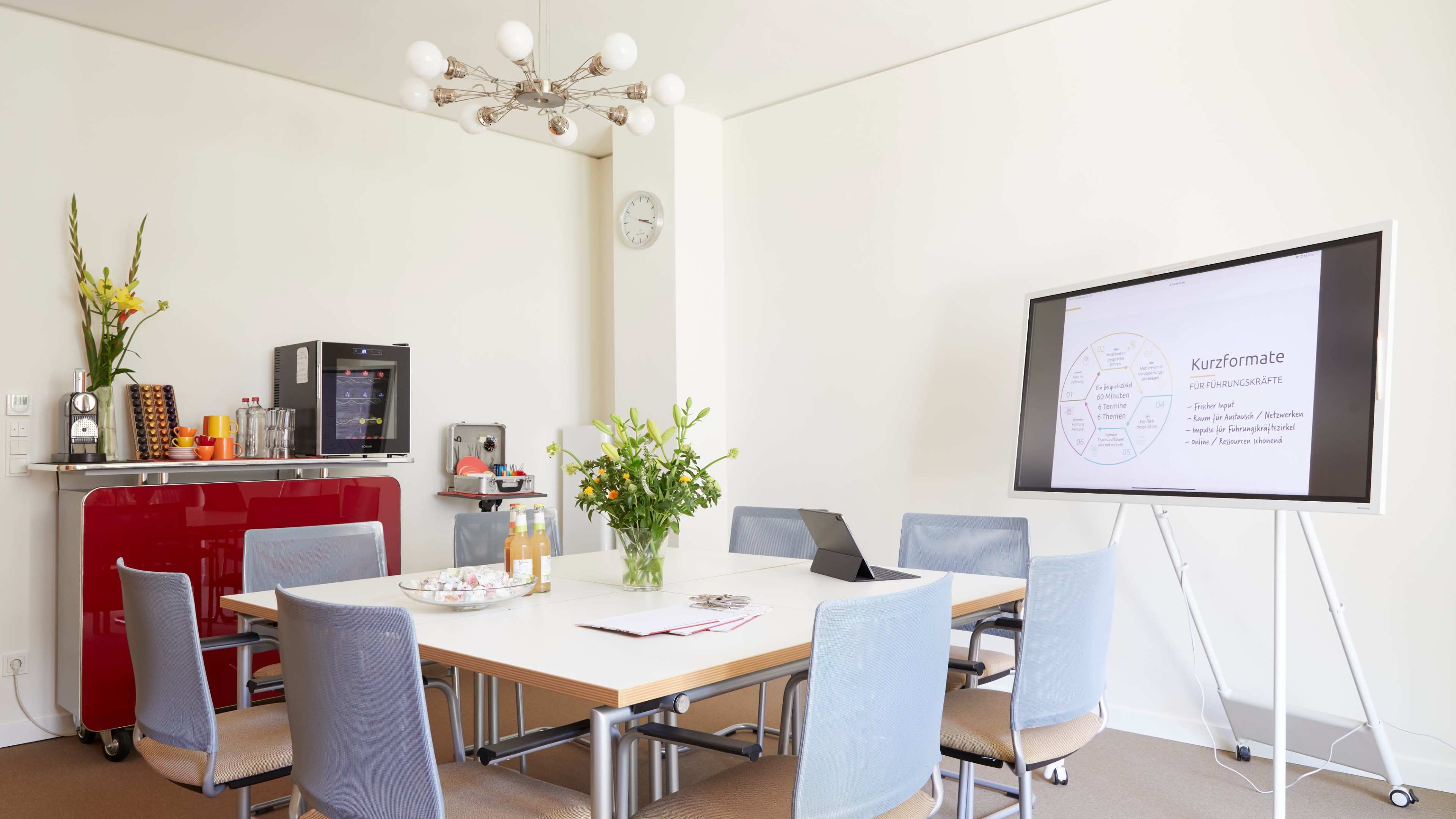 A modern meeting room features a large table surrounded by chairs, a display screen, and a small kitchenette with refreshments.